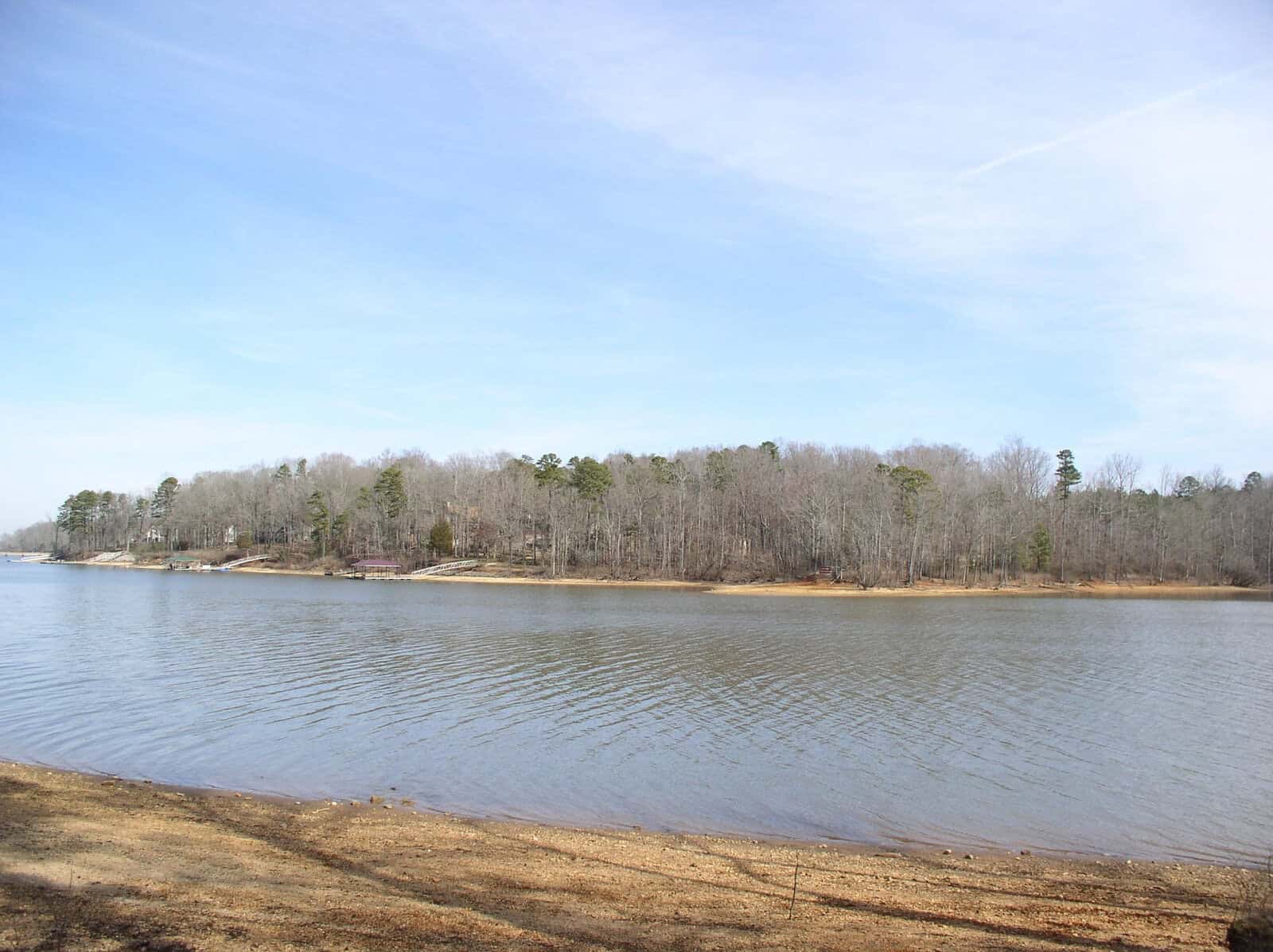 A calm lake with a sandy shore in the foreground and a forested area with scattered houses in the background under a partly cloudy sky.