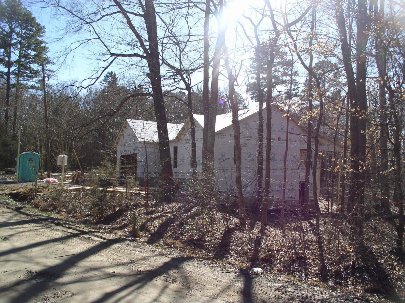 A construction site with a partially built house surrounded by bare trees under a clear sky, with a portable toilet on the left and construction materials scattered around.