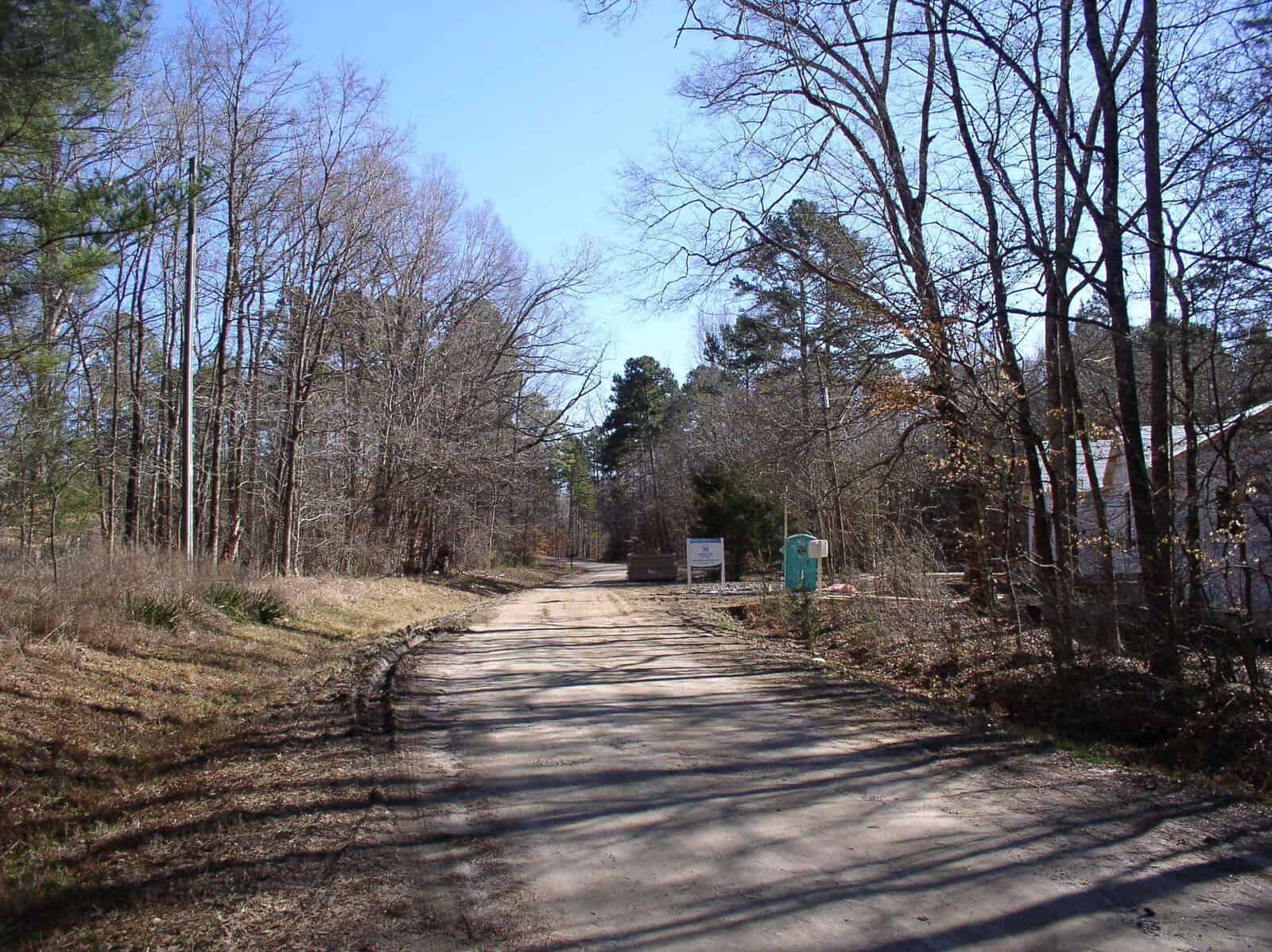 Dirt road leading through a wooded area with sparse leafless trees under a clear blue sky, with a small building and signage visible to the right.