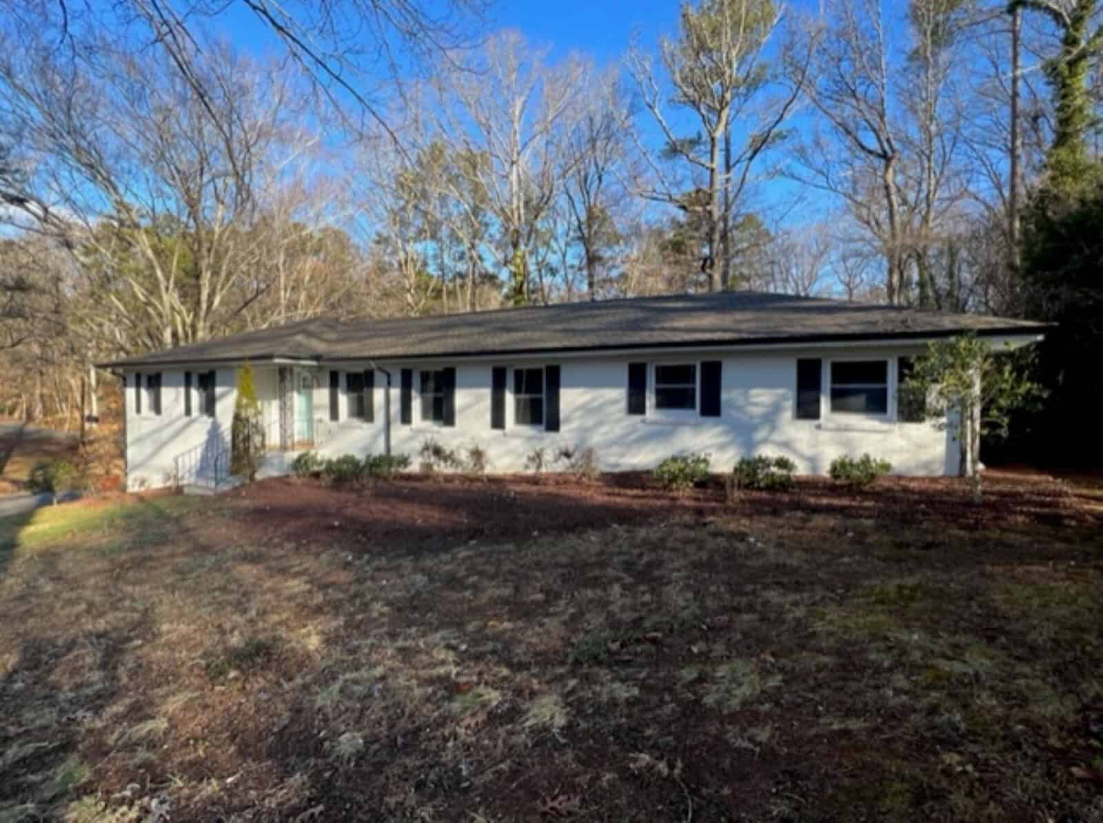 A single-story white house with a black roof, featuring a yellow front door, surrounded by a wooded area with some bare trees and a lawn with patches of grass, located at 374 Forest Drive,