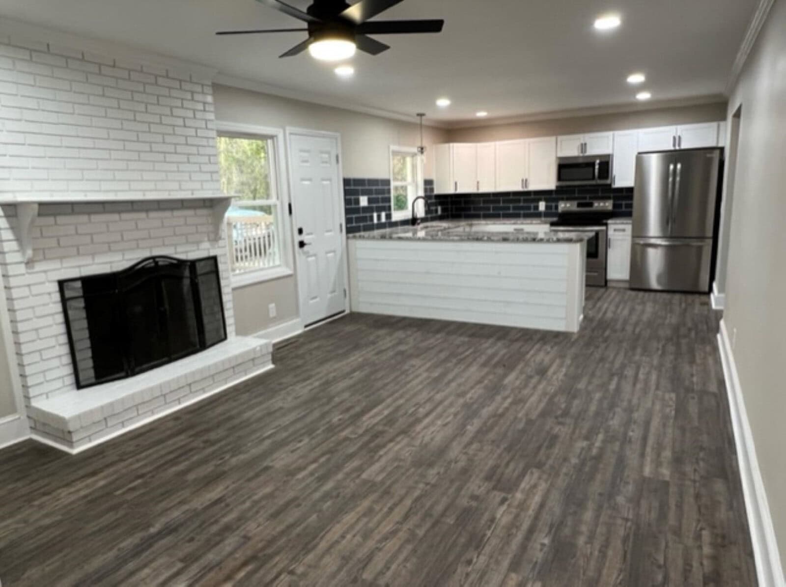 Modern kitchen with white cabinets, black countertops, and stainless steel appliances adjacent to a living area with a fireplace and hardwood flooring.