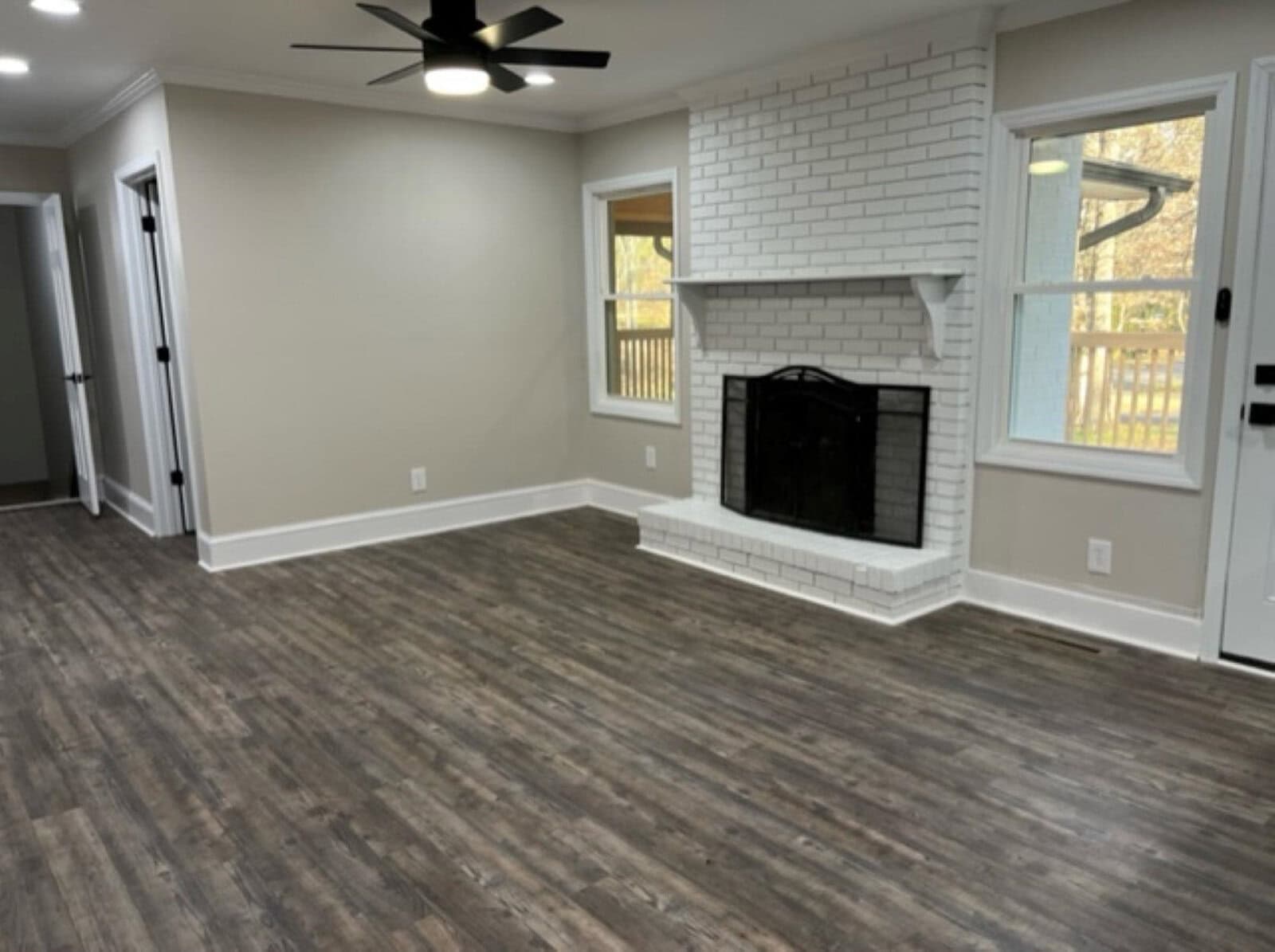 An empty room featuring a gray wood-style flooring, a white brick fireplace, and glass doors leading to an outside deck area, with a ceiling fan above.