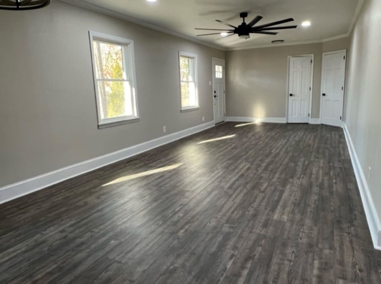 An empty room with light gray walls, dark wood flooring, windows with white trim, a ceiling fan, and closed doors.
