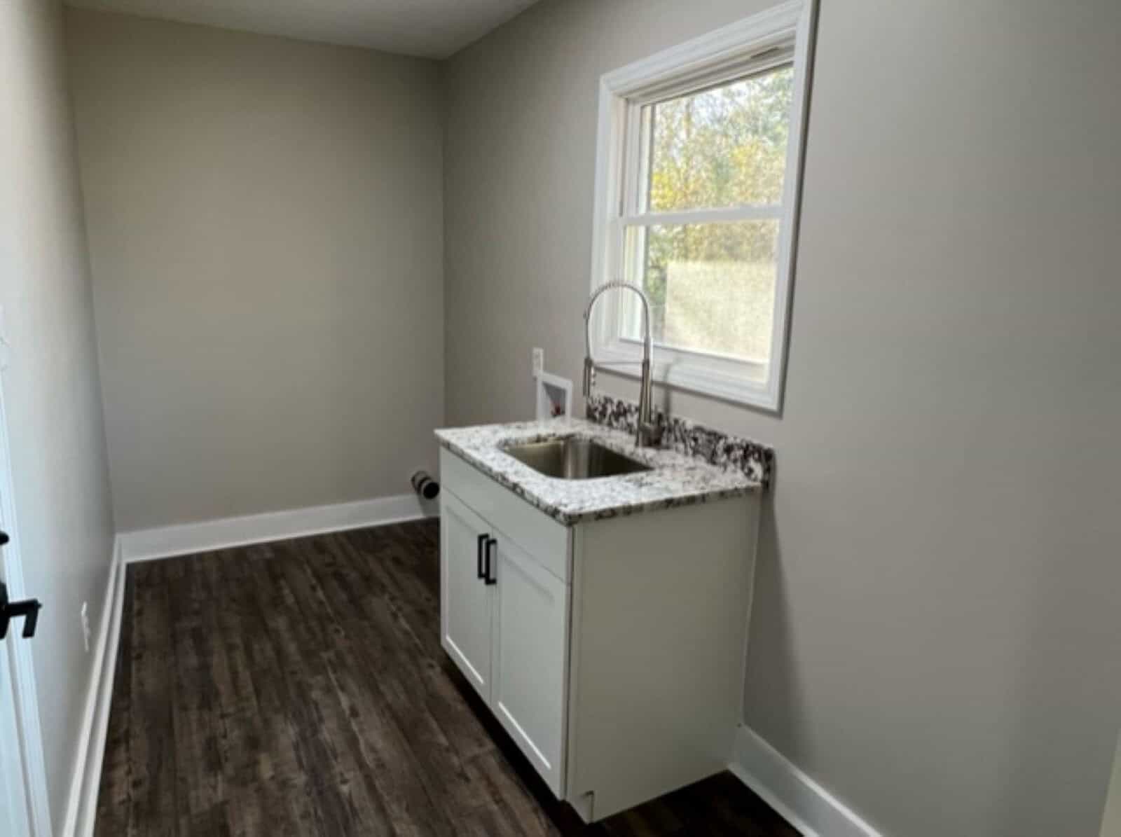A small, standalone kitchen sink cabinet is placed against a wall next to a window in a room with grey walls and wood flooring.