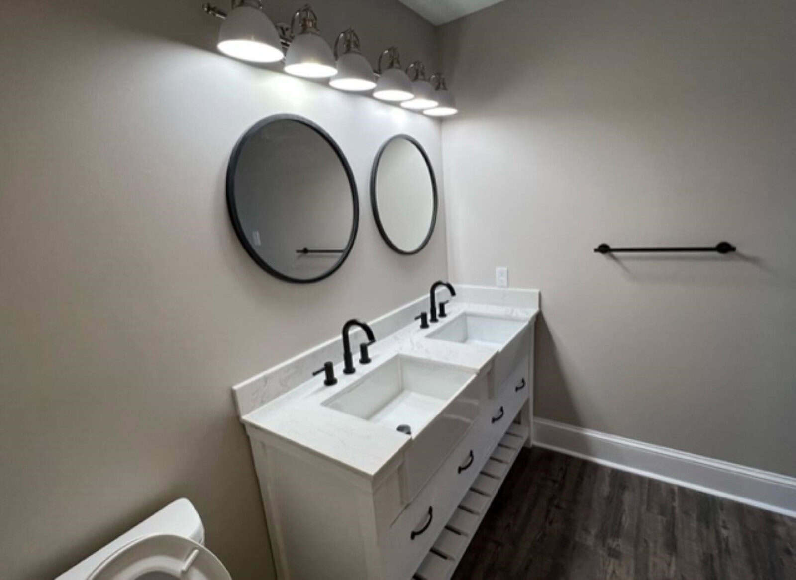 A modern bathroom featuring a white vanity with a marble countertop and two sinks, matte black faucets, two round mirrors above, a toilet to the left, and a towel bar on the right. the room has laminate flooring and overhead lighting.