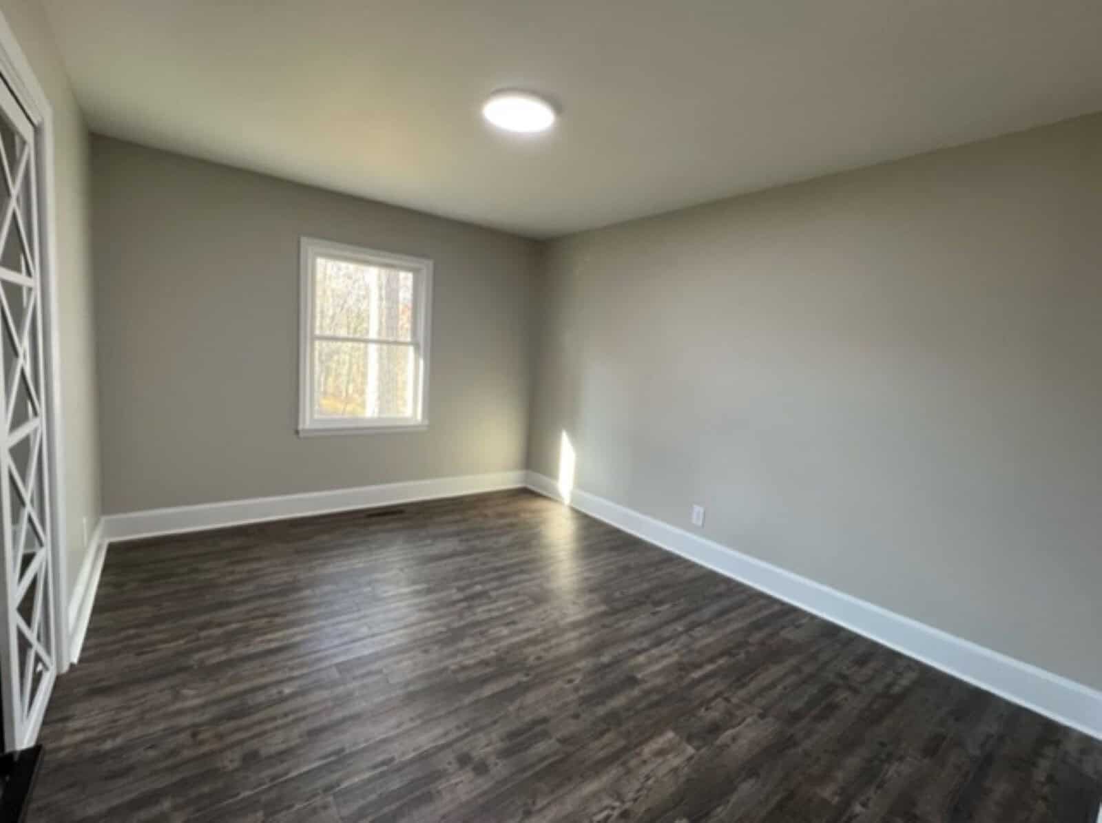 An empty room with gray walls, dark wood flooring, a single window with white trim, and one overhead light fixture.