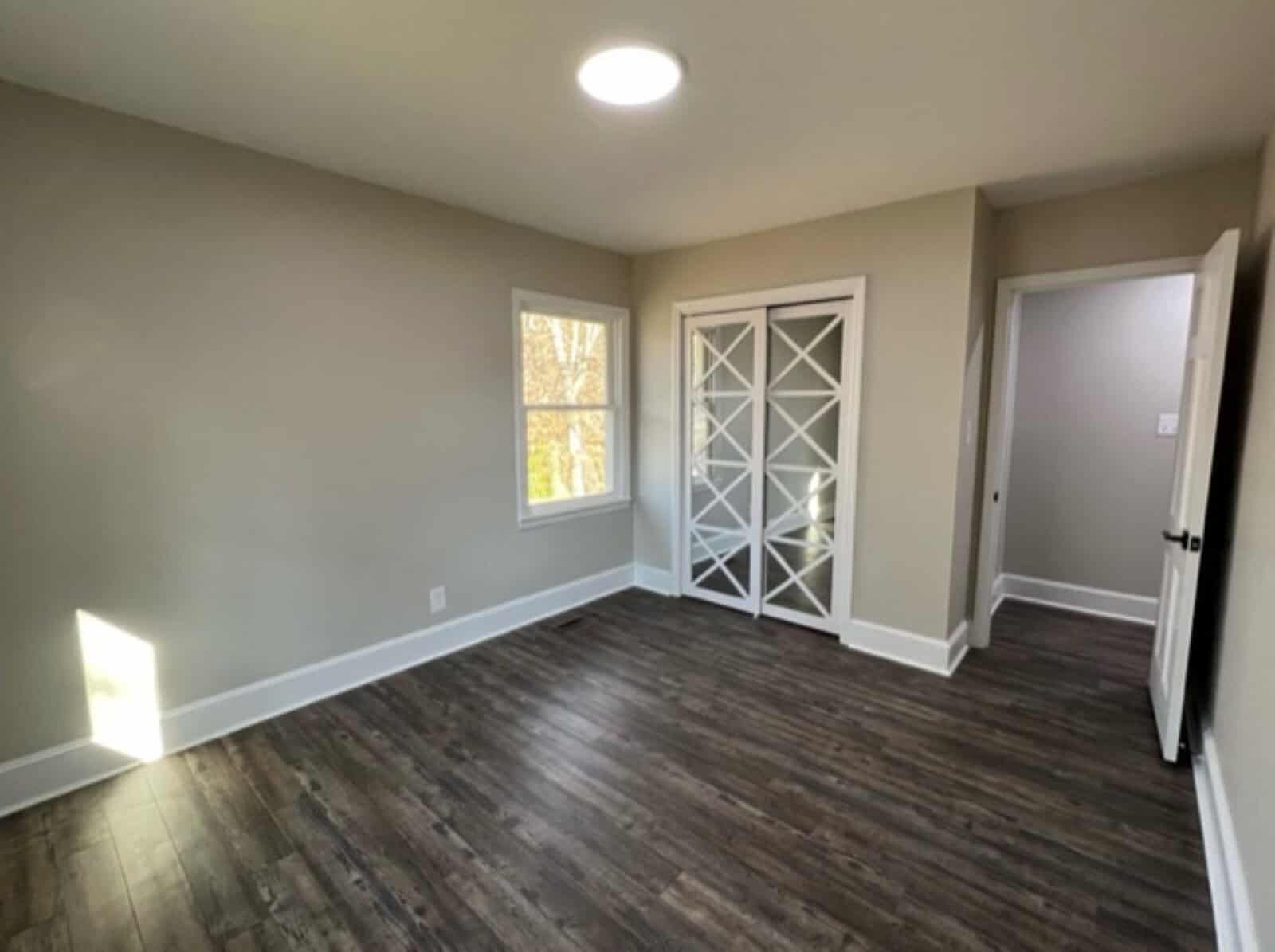 Spacious, empty room with natural light coming through a window, featuring laminate flooring, a white french door with diamond lattice patterns, and an open standard door.