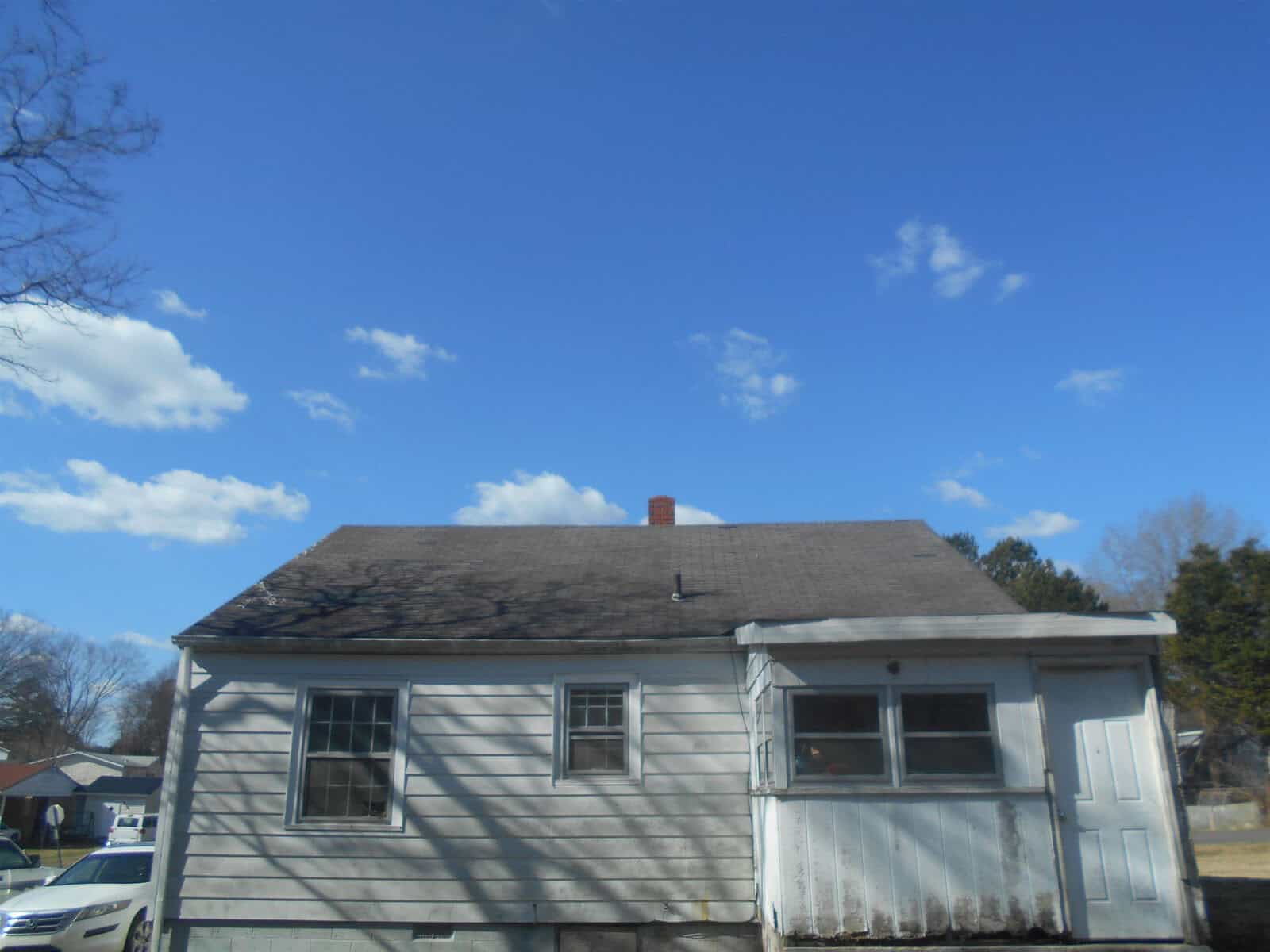 A single-story house with a shingled roof, featuring a small attached porch and a chimney, set against a clear blue sky.