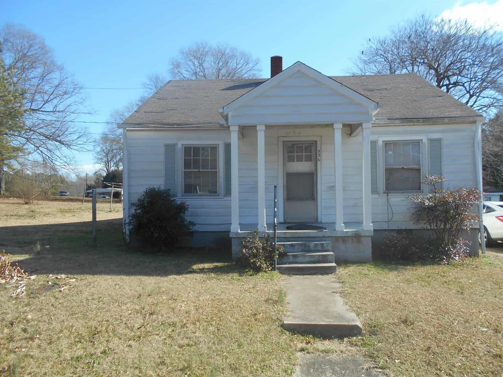 A small, single-story white house with a gabled roof and a central doorway flanked by two windows. the house appears to have a weathered exterior and is set on a plot of grass with a clear sky above and a car parked to the side.