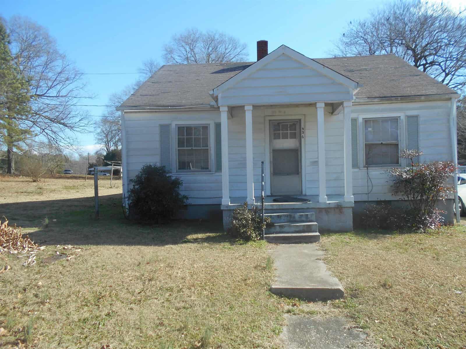 A modest single-story house with white siding, a gabled roof, and a central front door flanked by two windows, set on a plot with a lawn and bare shrubs, under a clear sky.