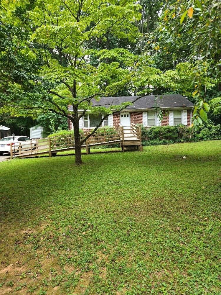 A modest single-story house with a white exterior and black shutters, featuring a small front porch, surrounded by a green lawn with trees and a partial wooden fence on one side.