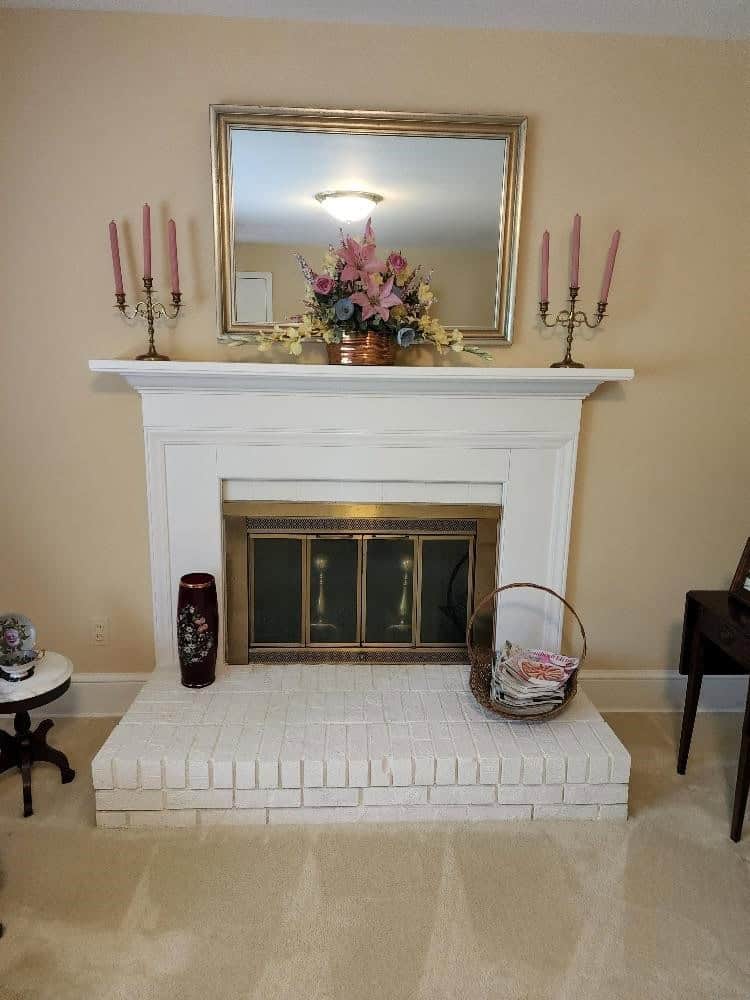 A traditional fireplace with a white mantel, decorated with a central floral arrangement, flanked by two pink candlesticks, beneath a gold-framed mirror, in a room with beige walls.