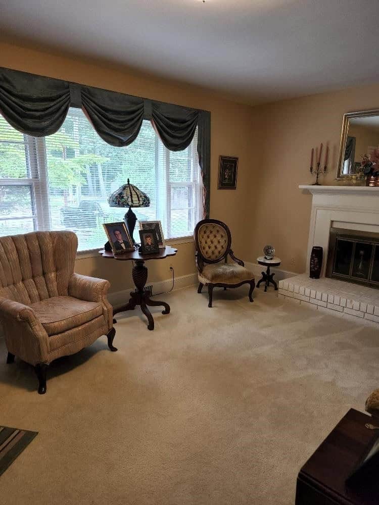 A cozy living room featuring a fireplace, a wingback chair, a round accent chair, and a side table with framed photographs, under a window with draped curtains.