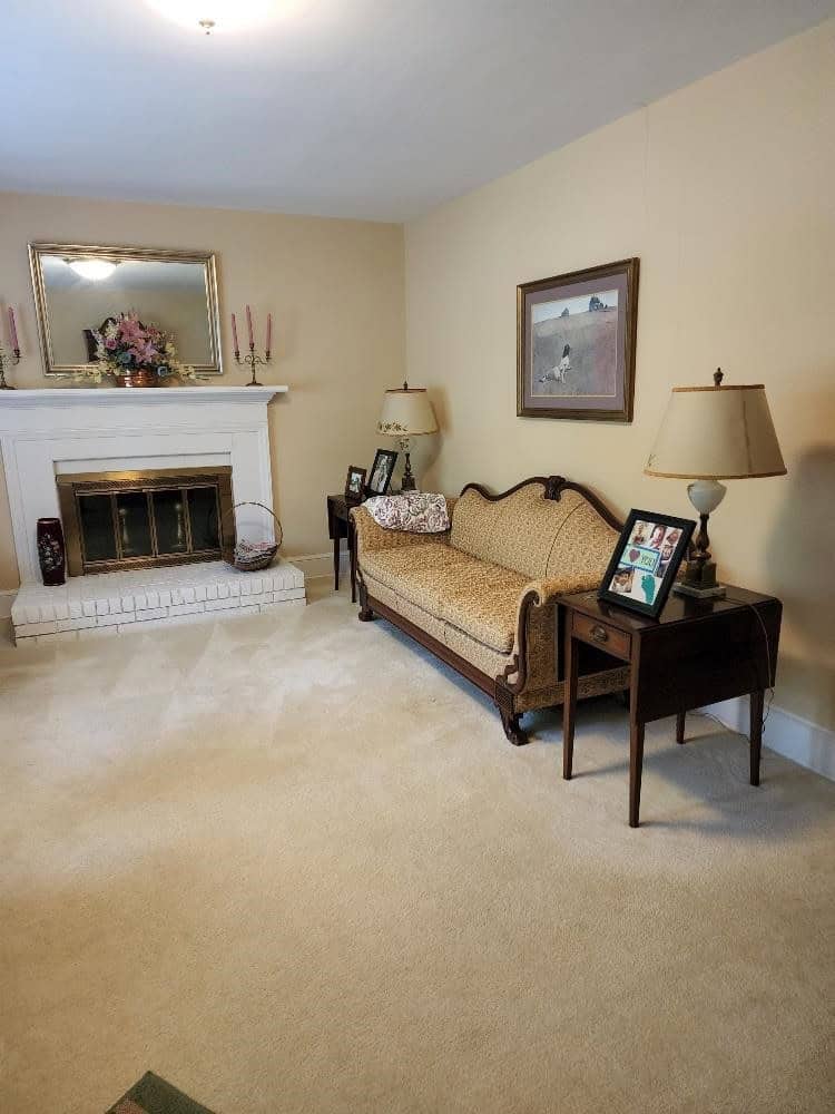 A traditional living room featuring a patterned sofa, a wooden side table with a lamp and photo frames, a fireplace with a decorative mirror above it, and a floral arrangement on the mantelpiece.
