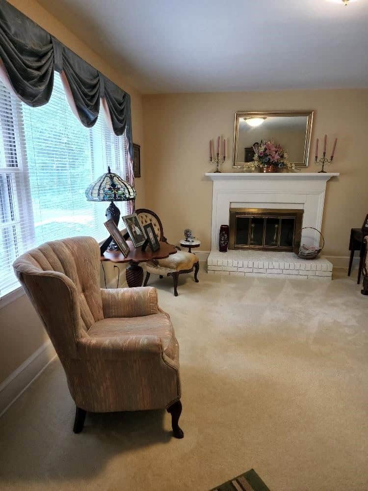 A living room with a single armchair, a small wooden table with a decorative lamp, a fireplace with a mirror and candles above it, and a window dressed with curtains.