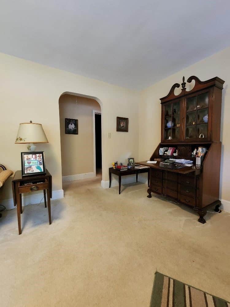 A living room with beige walls and carpet featuring a wooden secretary desk, a small table with a lamp, and a chair by the wall. an arched doorway is visible on the left.