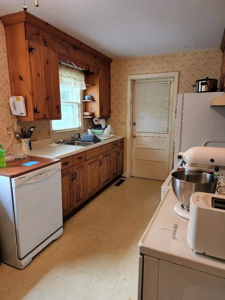 A dated kitchen with wooden cabinets, beige countertop, and older appliances, including a white dishwasher, refrigerator, stand mixer, and a microwave.