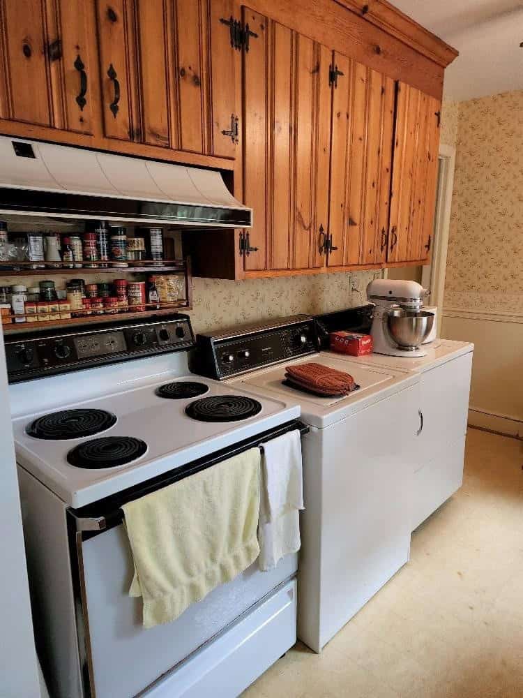 A well-used kitchen with wooden cabinets, an electric stove with a dishtowel hanging from its handle, and a countertop cluttered with various spices and a kitchen mixer.
