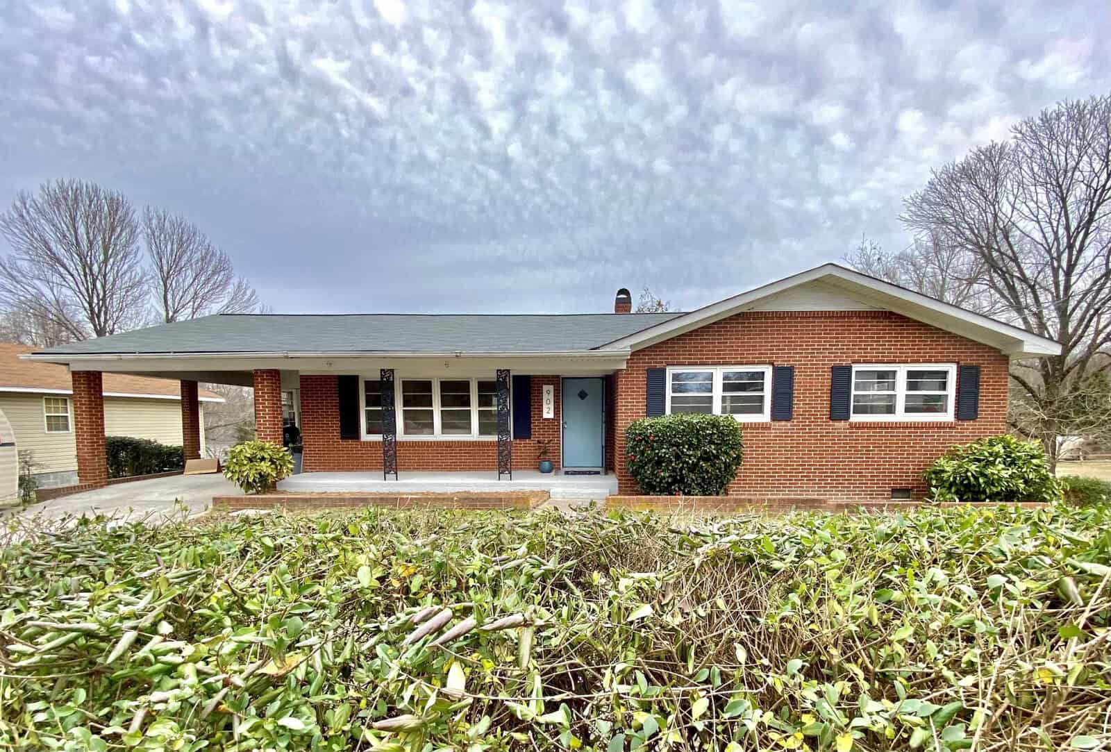 A single-story brick house with a carport, located at 902 Bridgers Street Henderson NC, featuring a blue front door and surrounded by a yard with overgrown vegetation.