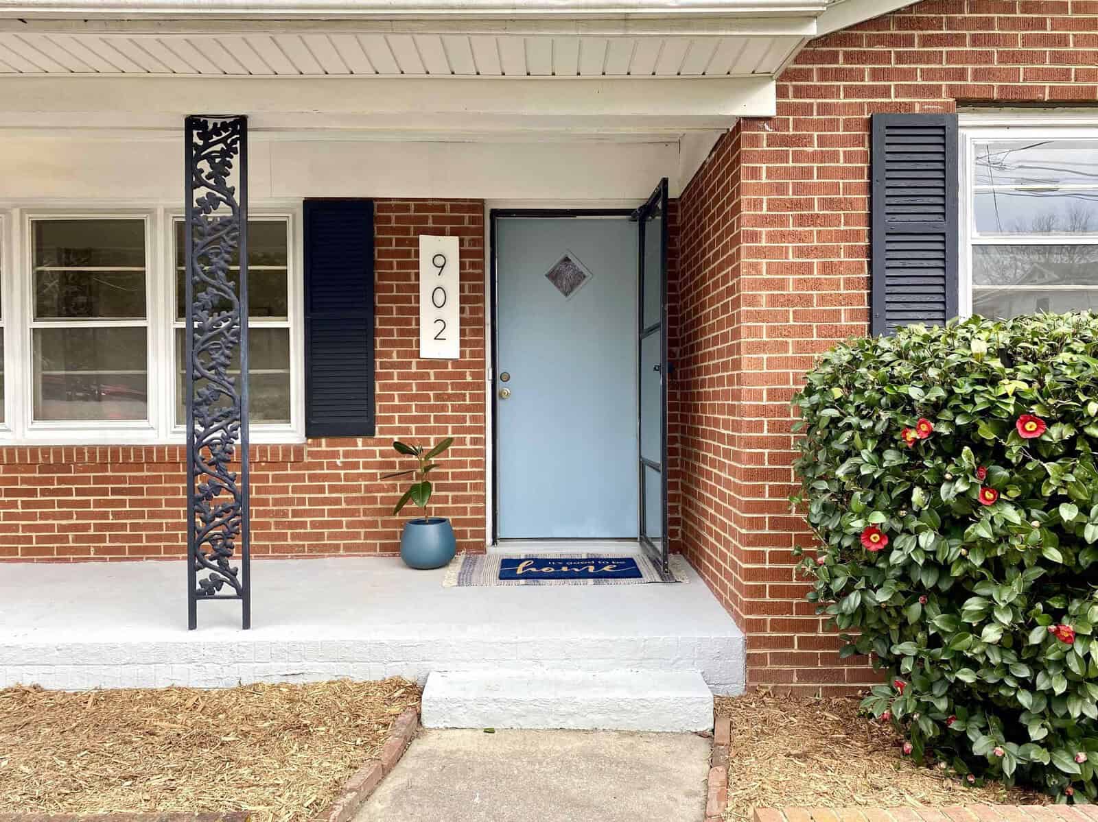 A residential front entrance featuring a blue door with the house number 902, flanked by red-brick walls, a white-framed window to the left, and a bush with red flowers to the right. a doormat lies in front of the door and a decorative plant is situated in a blue pot on the porch.