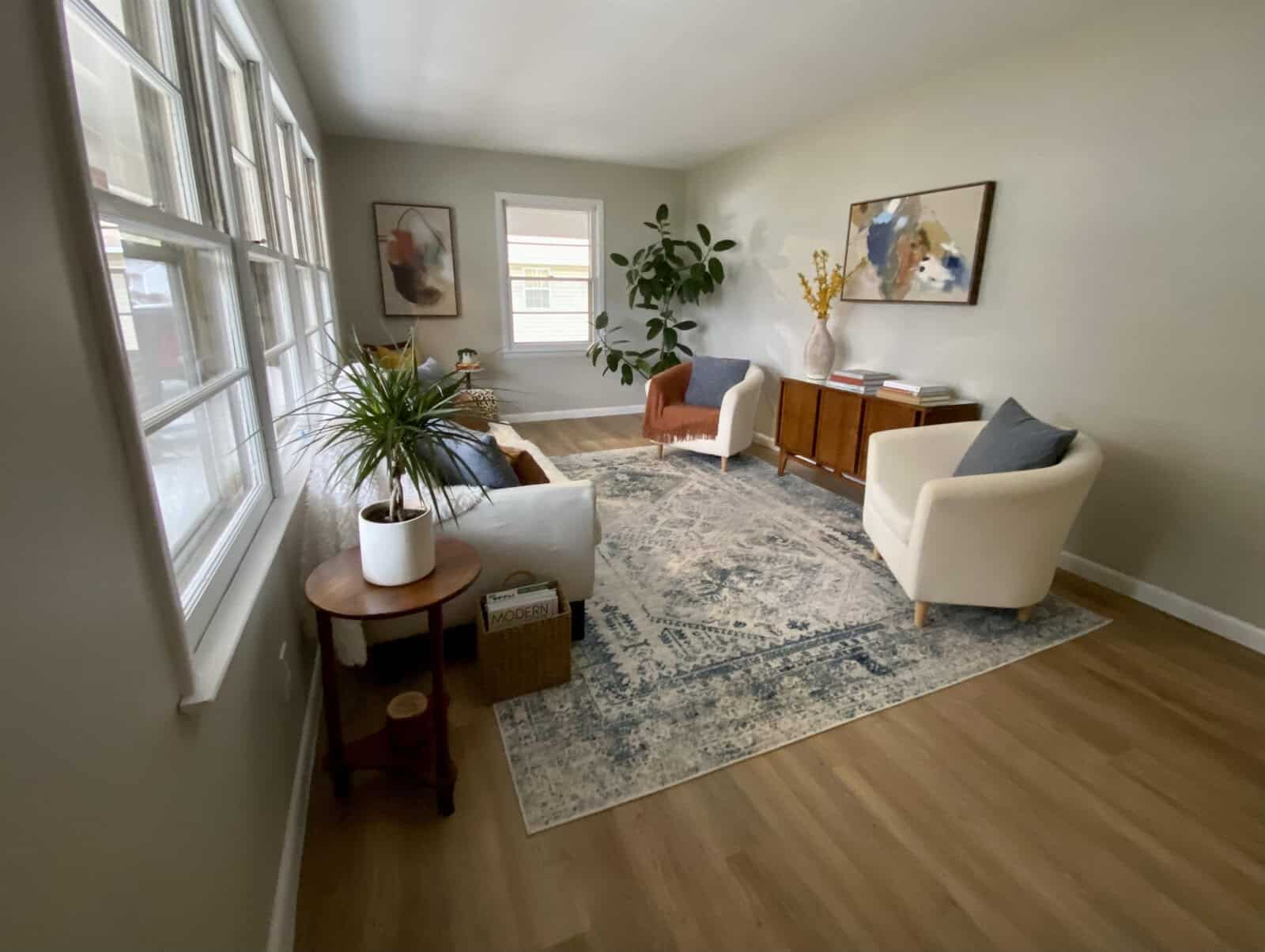 A neatly arranged living room with contemporary furniture, including two sofas, a central coffee table, a side table with a plant, and a credenza against the wall. the space is accented by a large area rug, decorative plants, and framed artwork on the walls. large windows allow for ample natural light.