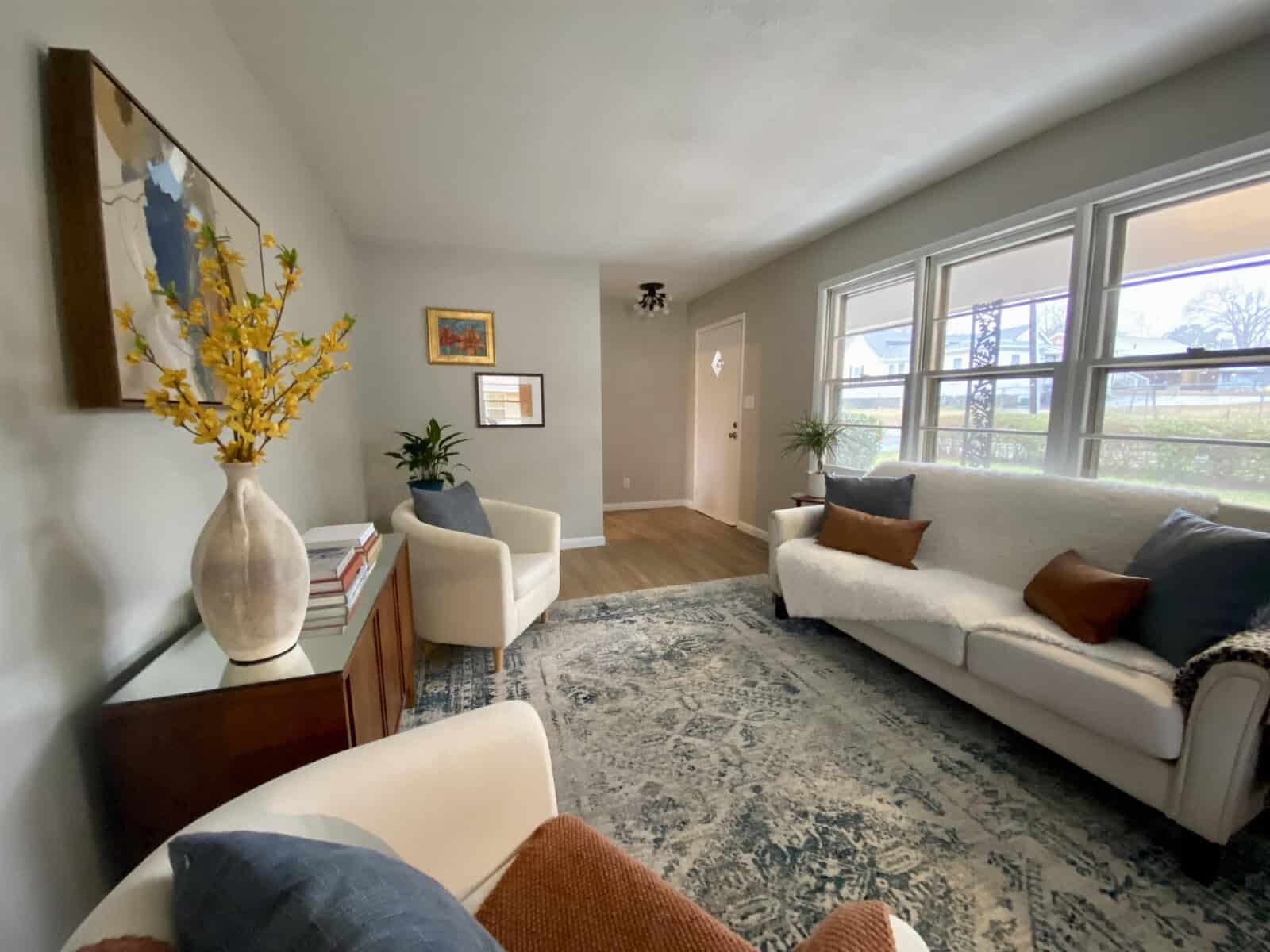 A neatly arranged living room with a white sofa, accent chairs, a blue area rug, and decorative elements like wall art and vases, with ample natural light coming in through large windows.