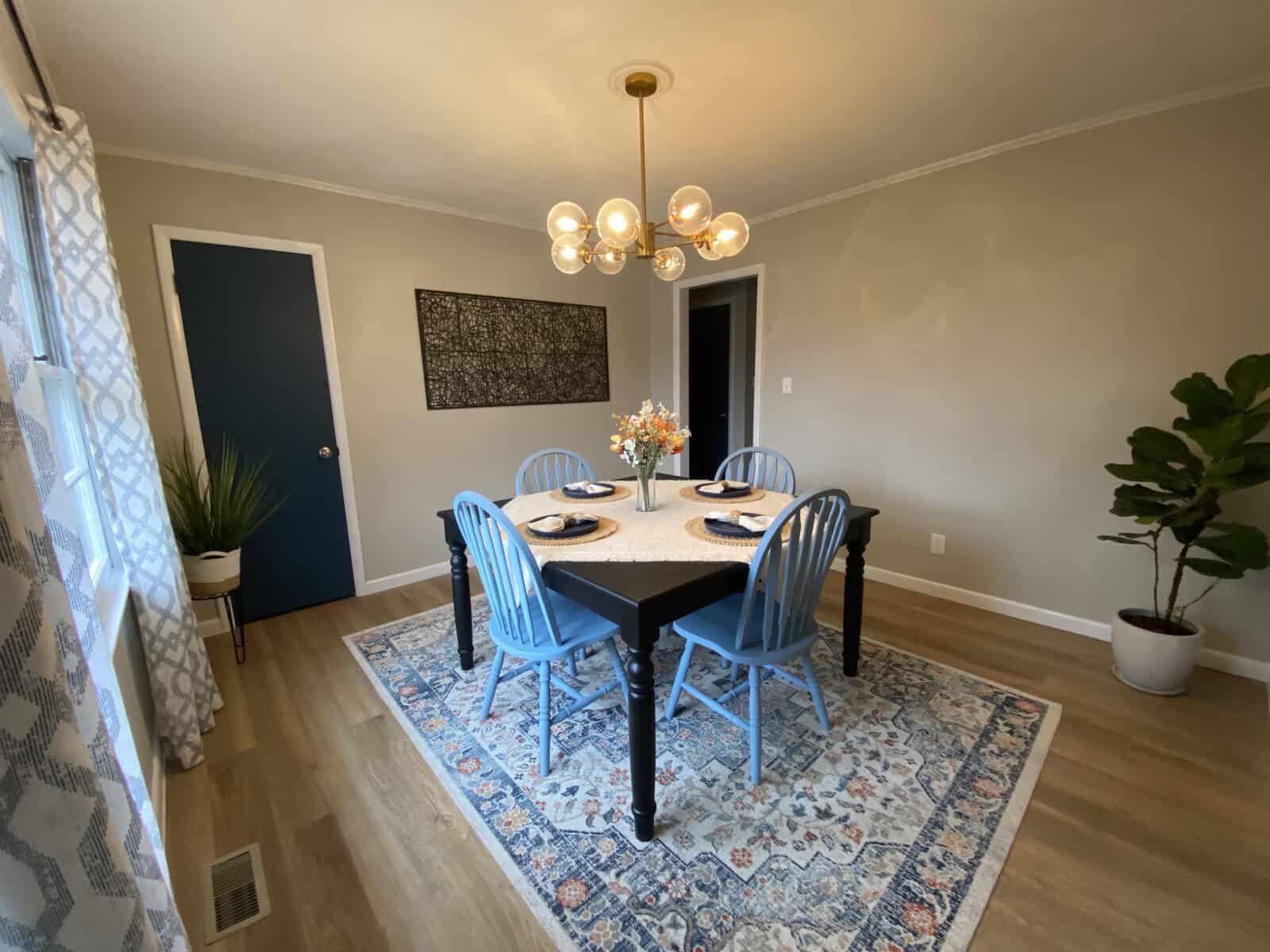 A well-lit dining room featuring a round table set for four with blue chairs, a vibrant area rug, a modern chandelier above, and plants adding a touch of greenery.