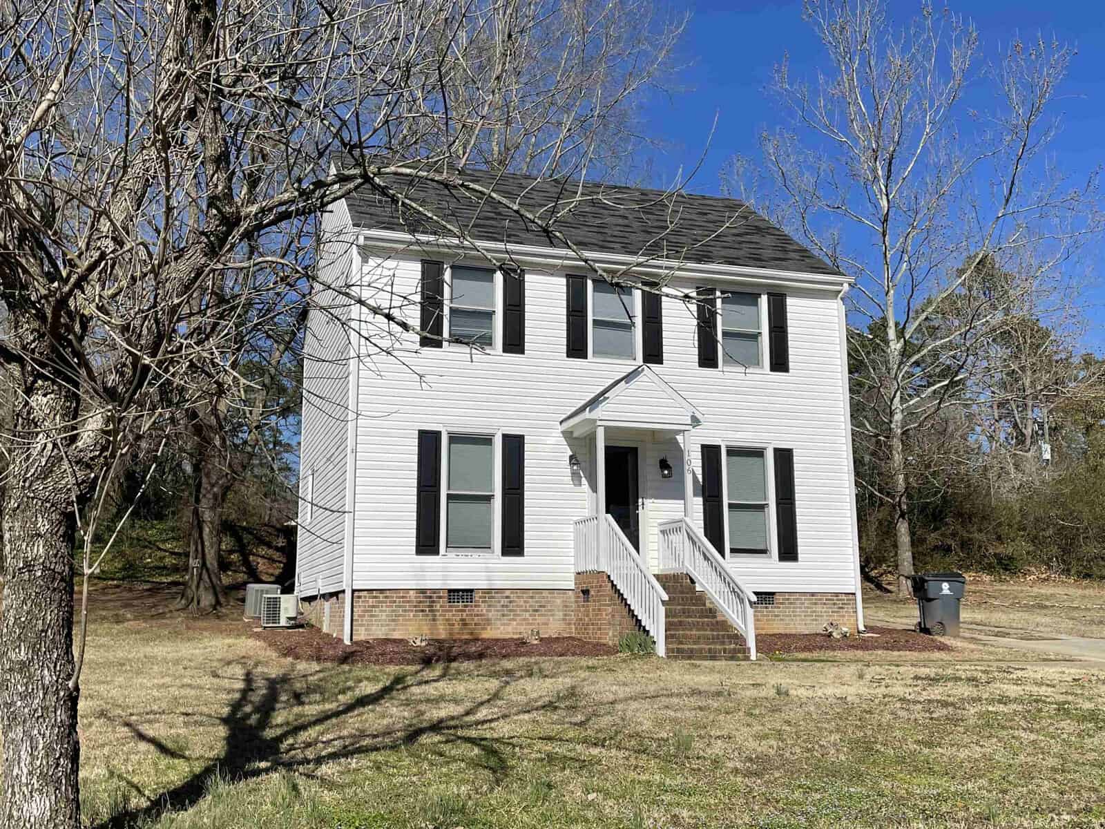 A two-story white house with black shutters, featuring a central front door with steps leading up to it, flanked by two ground-floor windows and a second-story balcony with three windows. there is a bare tree on the left and a blue recycling bin at the curb in front of the house. the sky is clear and it appears to be daytime.