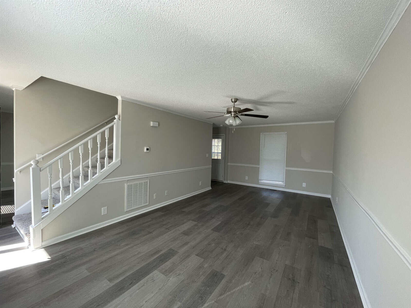 An empty, well-lit room featuring grey flooring, a staircase with a white railing on the left, a ceiling fan, and neutral colored walls.