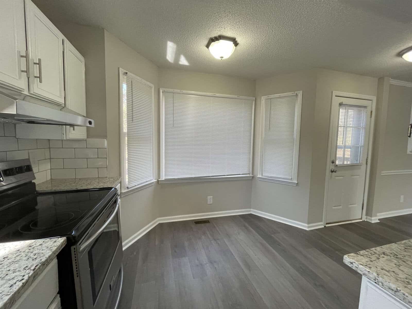 A well-lit, empty kitchen with white cabinetry, modern appliances, and a gray laminate flooring. three windows with closed blinds are centered on the far wall.