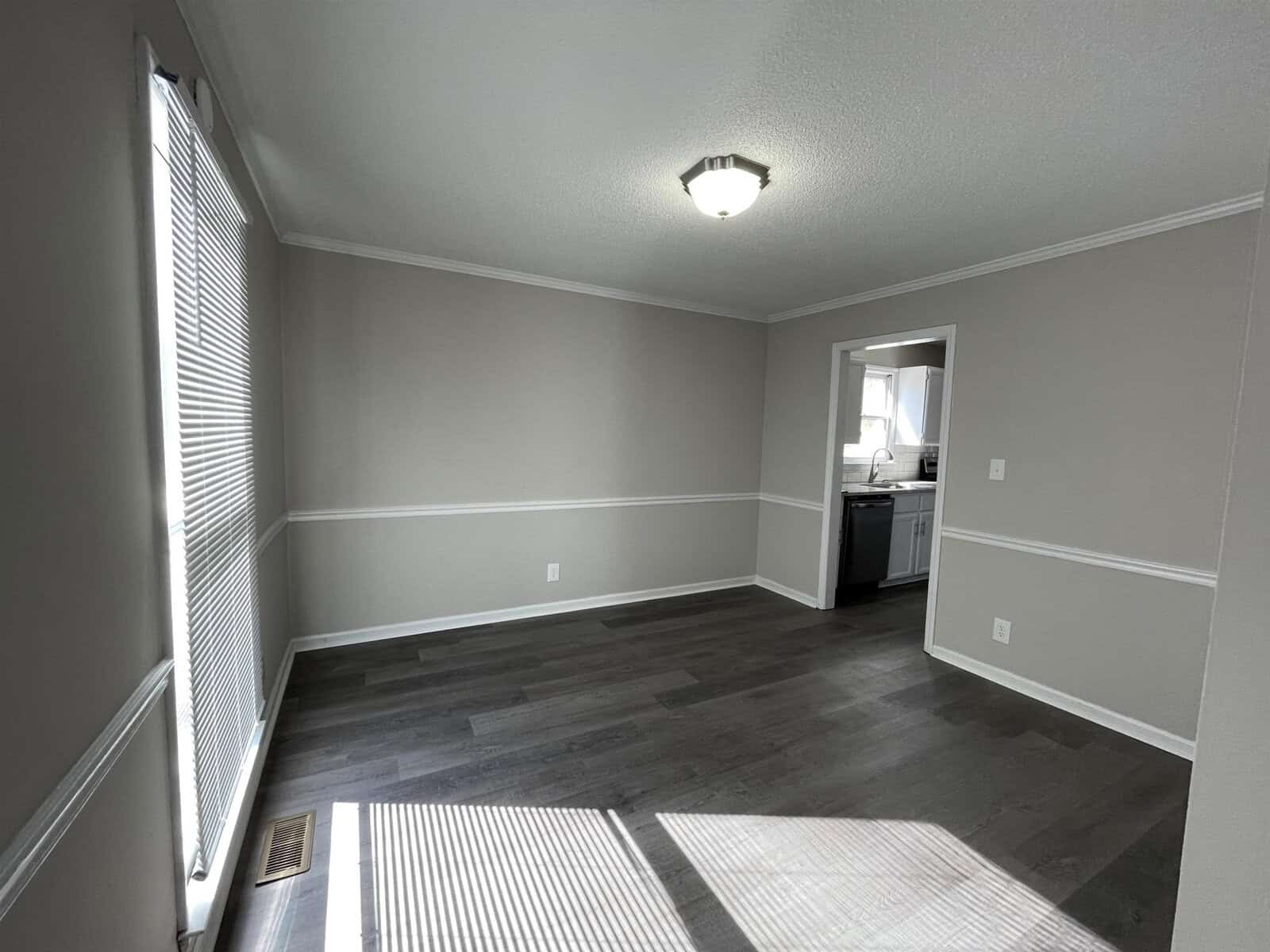 A clean, empty room with gray flooring and white walls featuring a chair rail, one window with blinds partially drawn, and an open doorway leading to a kitchen area. sunlight is casting shadows on the floor.