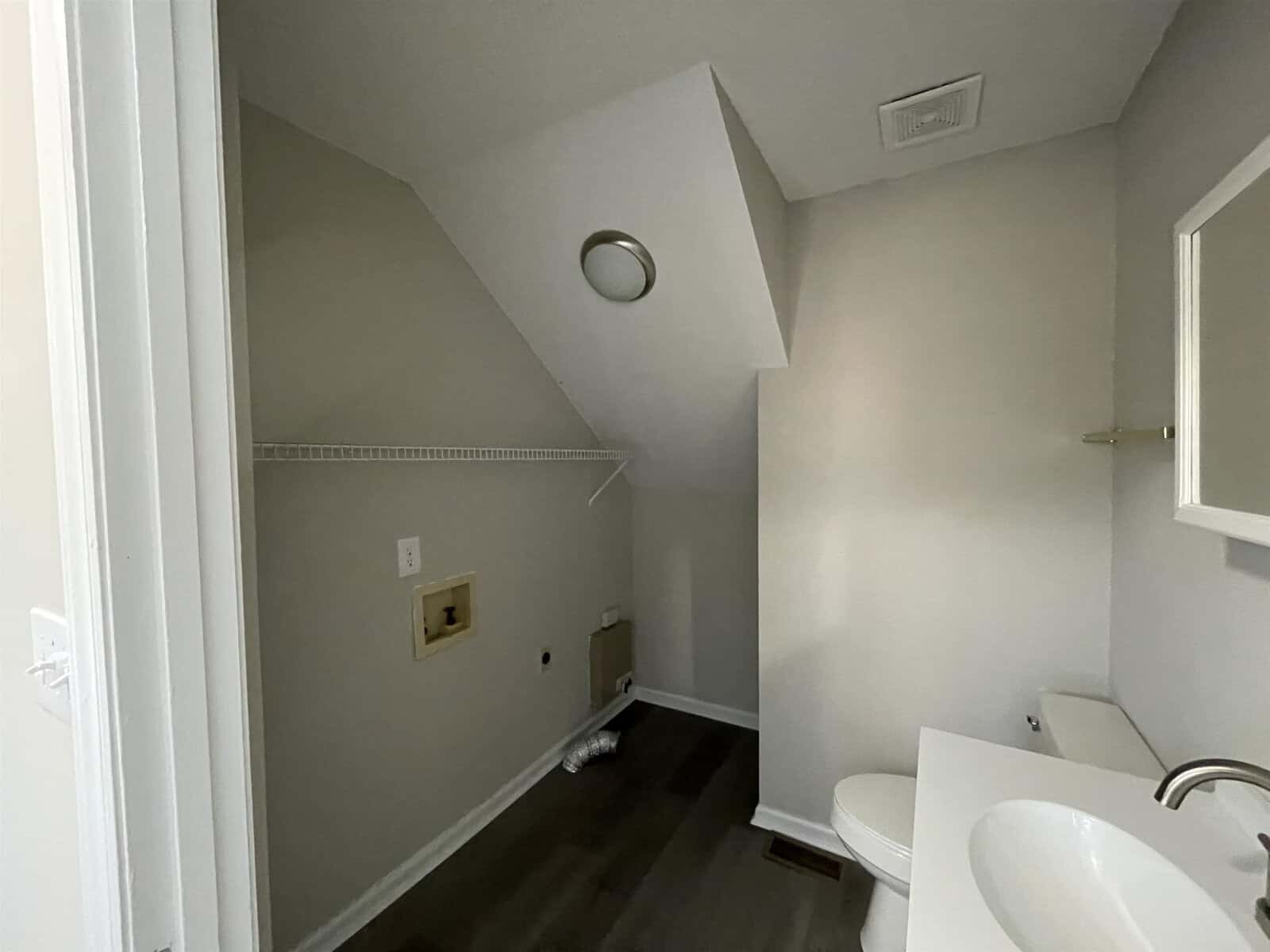 An empty, small, and narrow laundry room with white walls, featuring a washer hook-up and venting, along with shelving above, and a toilet to the right side.