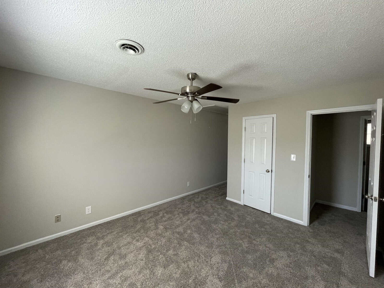 An empty room with light gray walls, carpeted flooring, a ceiling fan with lights, and a closed white door on the right wall. there is an open doorway to another room on the left.