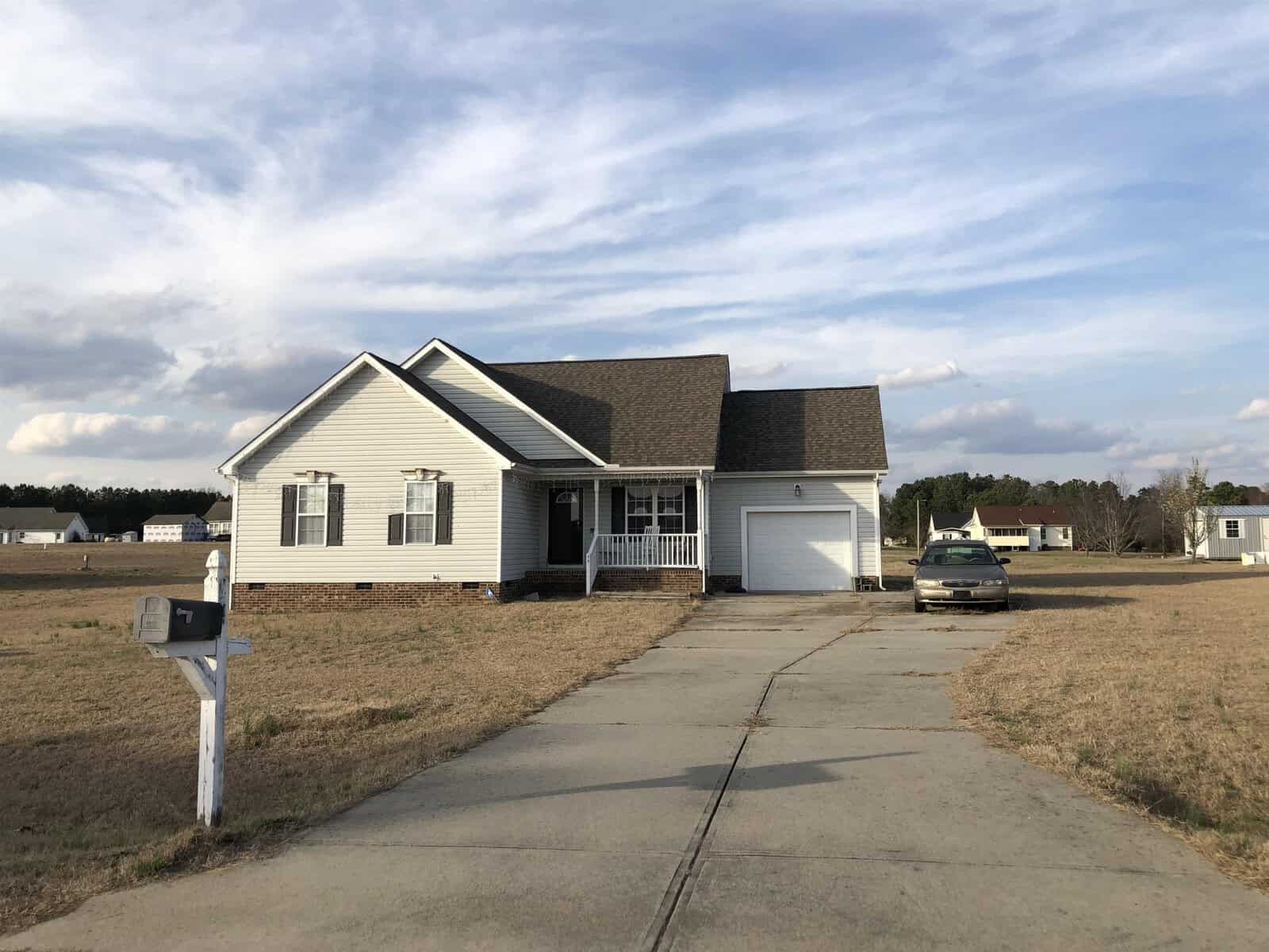 A single-story white house with a gabled roof, featuring an attached garage, a front porch, and a driveway leading to a mailbox, set against a partly cloudy sky. a sedan is parked in the driveway.