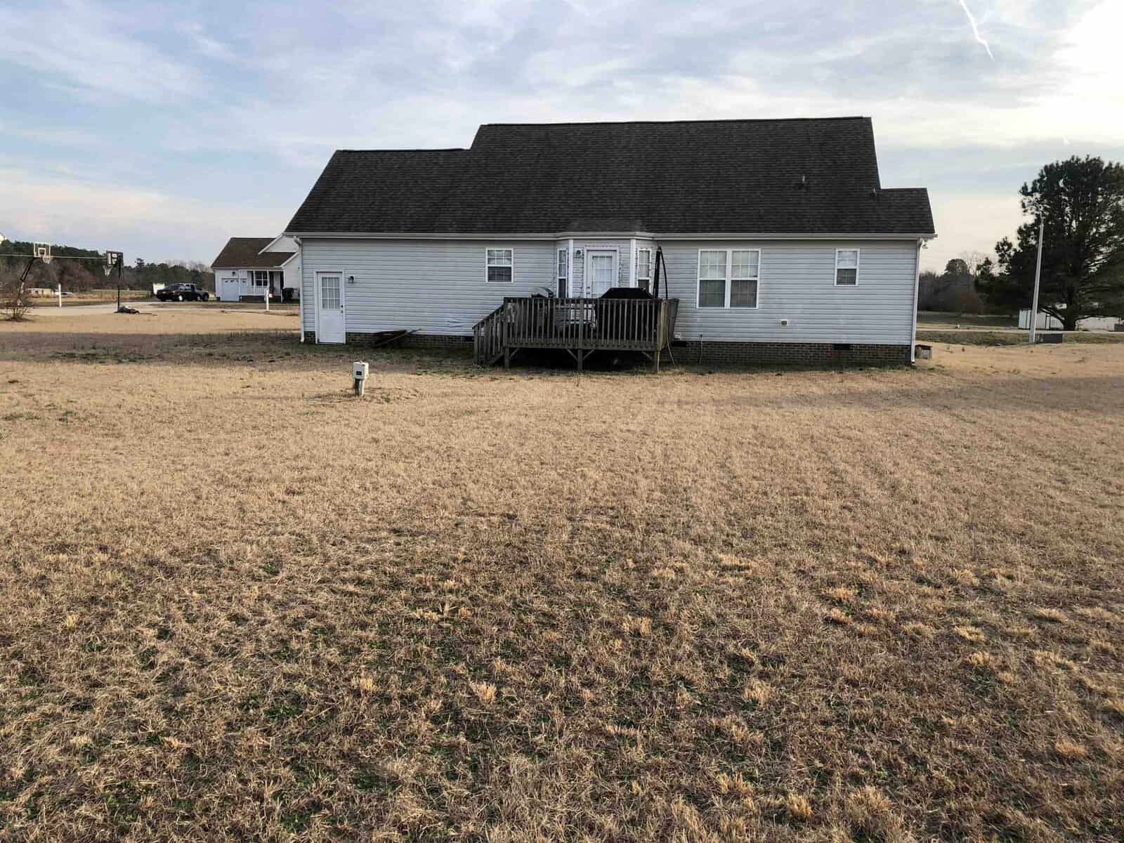 A single-story house with a wooden deck located in a wide, open field with dry grass, under a partly cloudy sky.
