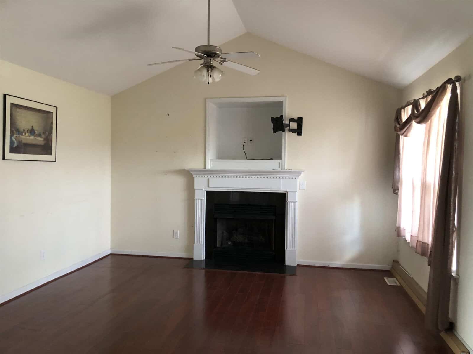 A spacious room featuring hardwood floors, a white fireplace with a mantel, a ceiling fan, and a wall-mounted television bracket. there is minimal decoration with one framed picture on the wall and curtains on a window to the right.