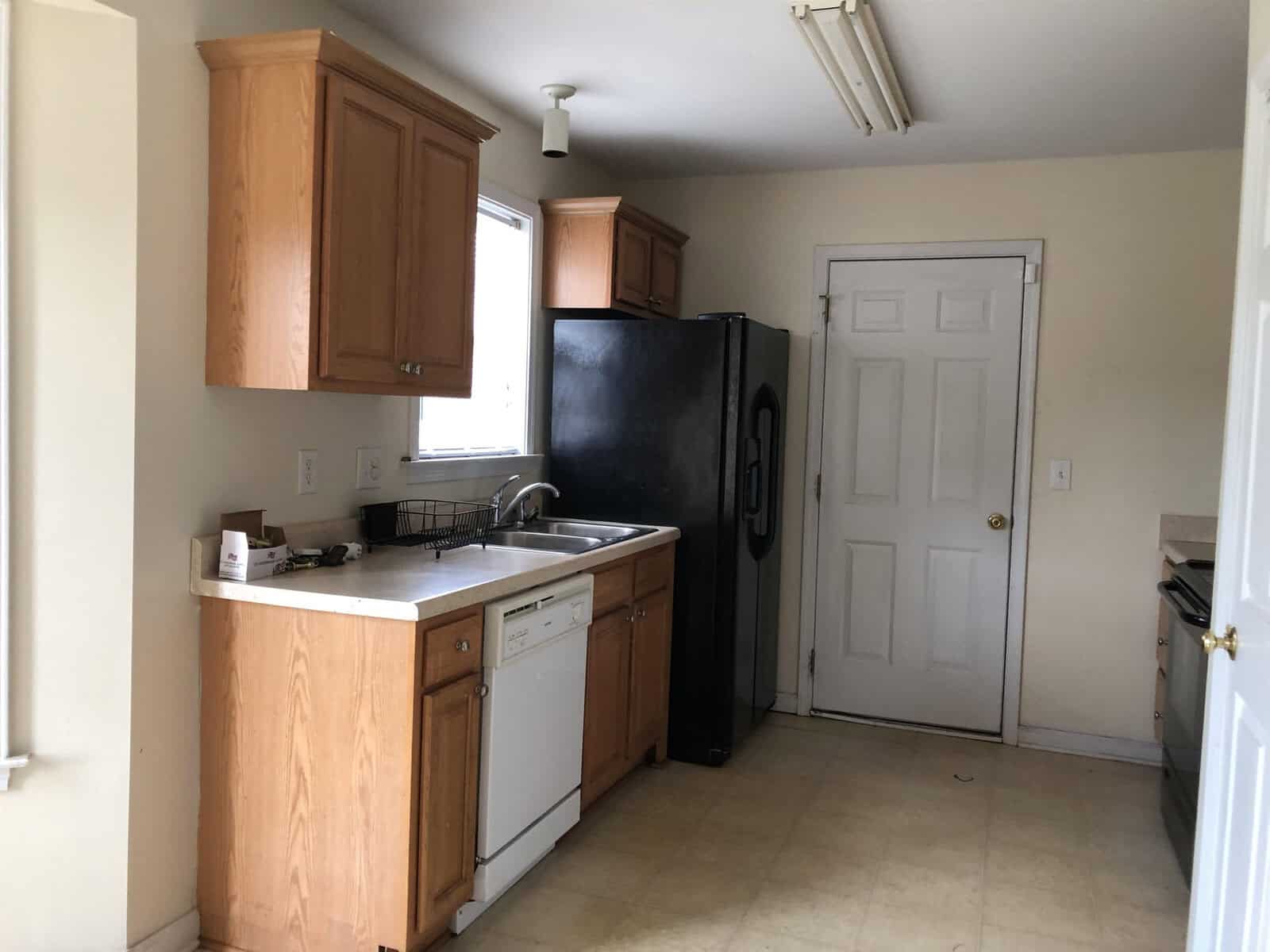 A modest kitchen with wooden cabinets, a white dishwasher to the left of the sink, a black refrigerator to the right, and a door at the end of the room. the kitchen appears to have minimal appliances and is lit by natural light.