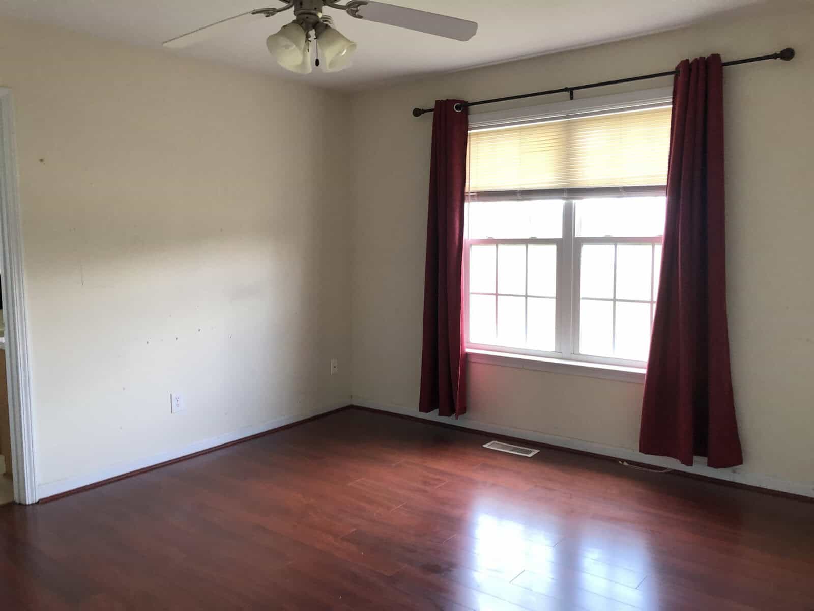 Empty room with wooden flooring, a window with red curtains, and a ceiling fan.