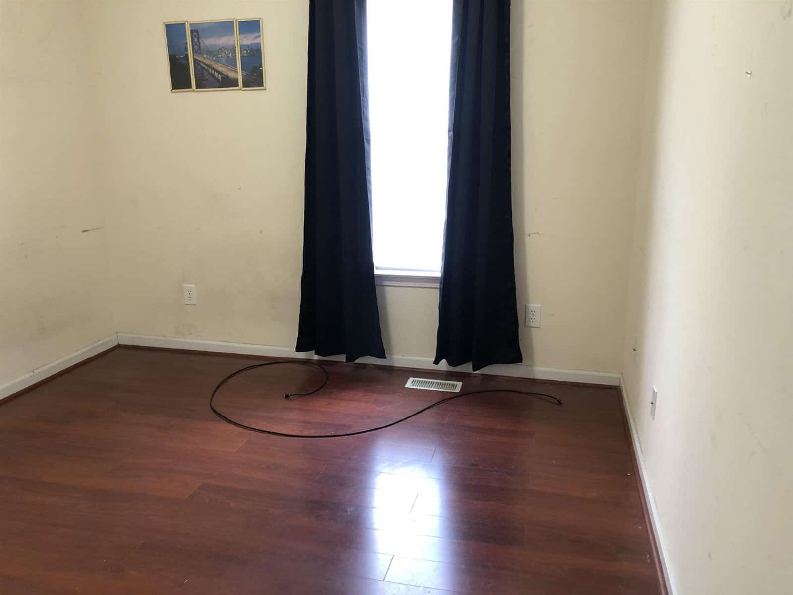 An empty room with hardwood floors, black curtains on a window, a single wall-mounted picture frame, and a cable on the floor.