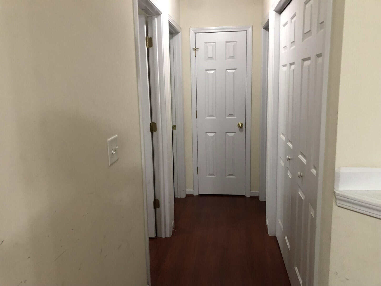 A hallway with white walls and three closed white doors, featuring wooden flooring and a visible light switch on the left wall.