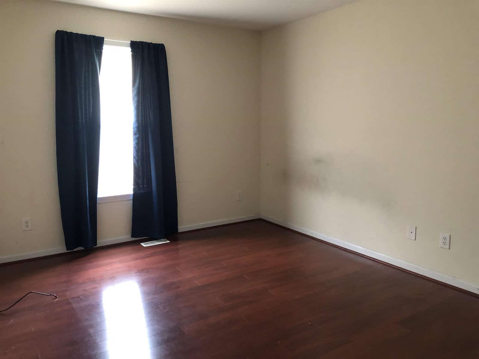 An empty room with beige walls, hardwood flooring, a window with dark curtains, and a visible scuff mark on the wall.