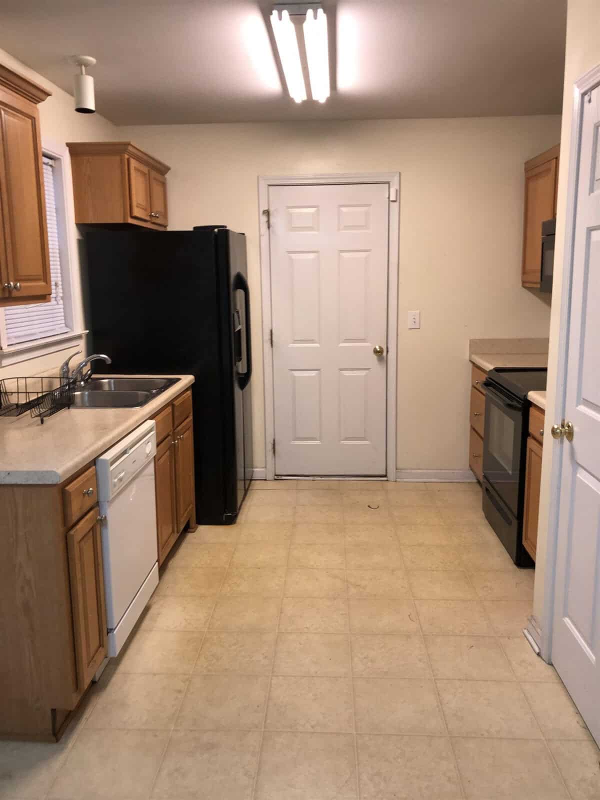 A domestic kitchen featuring wood cabinetry, a black refrigerator, and white appliances, with a tiled floor leading to a closed white door.