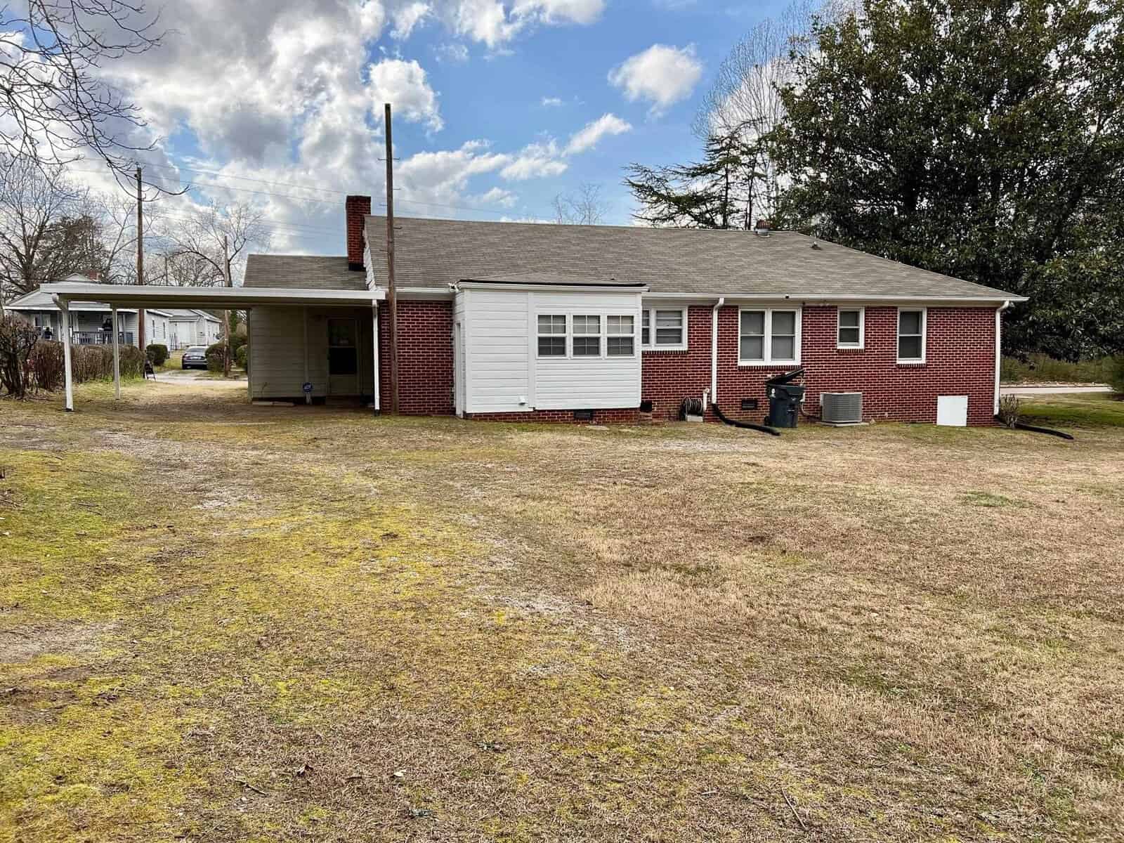 A single-story brick house with a white front porch, a bay window, a prominent chimney, an attached carport, and a lawn in need of maintenance.