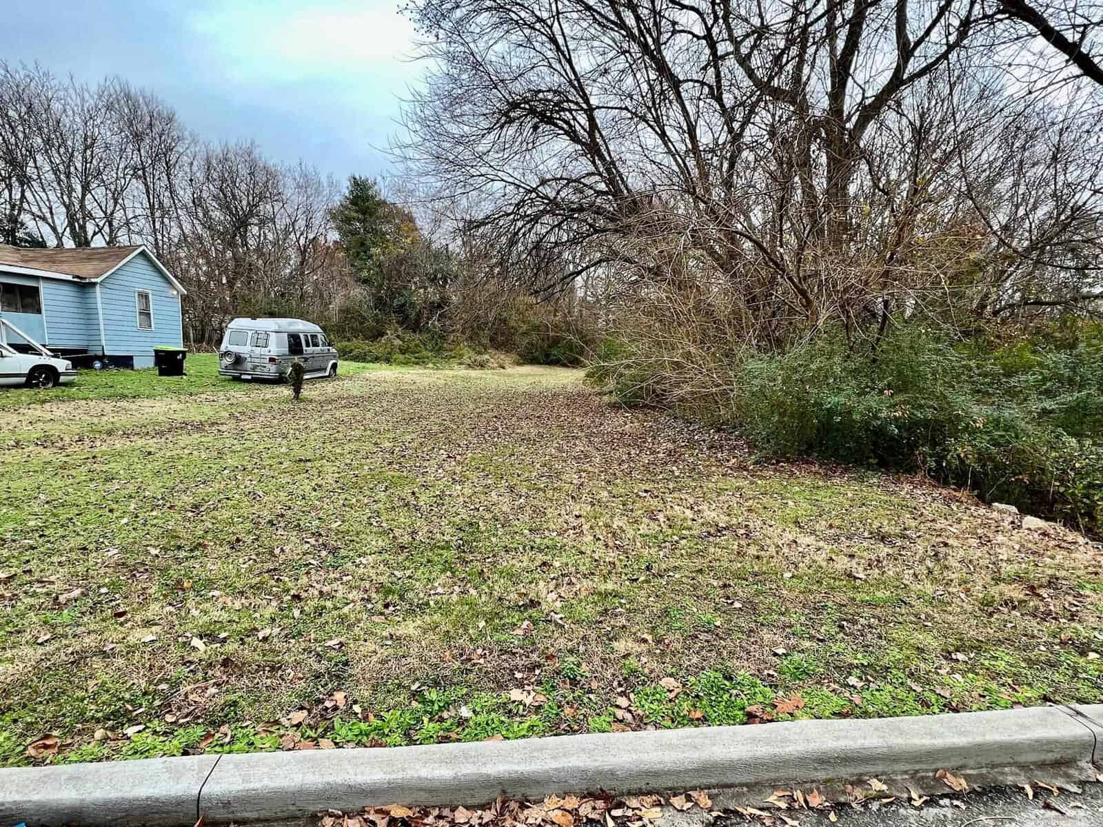 A residential area featuring a grassy field with leafless trees in the background, a blue house to the left, and a white van parked in the driveway.