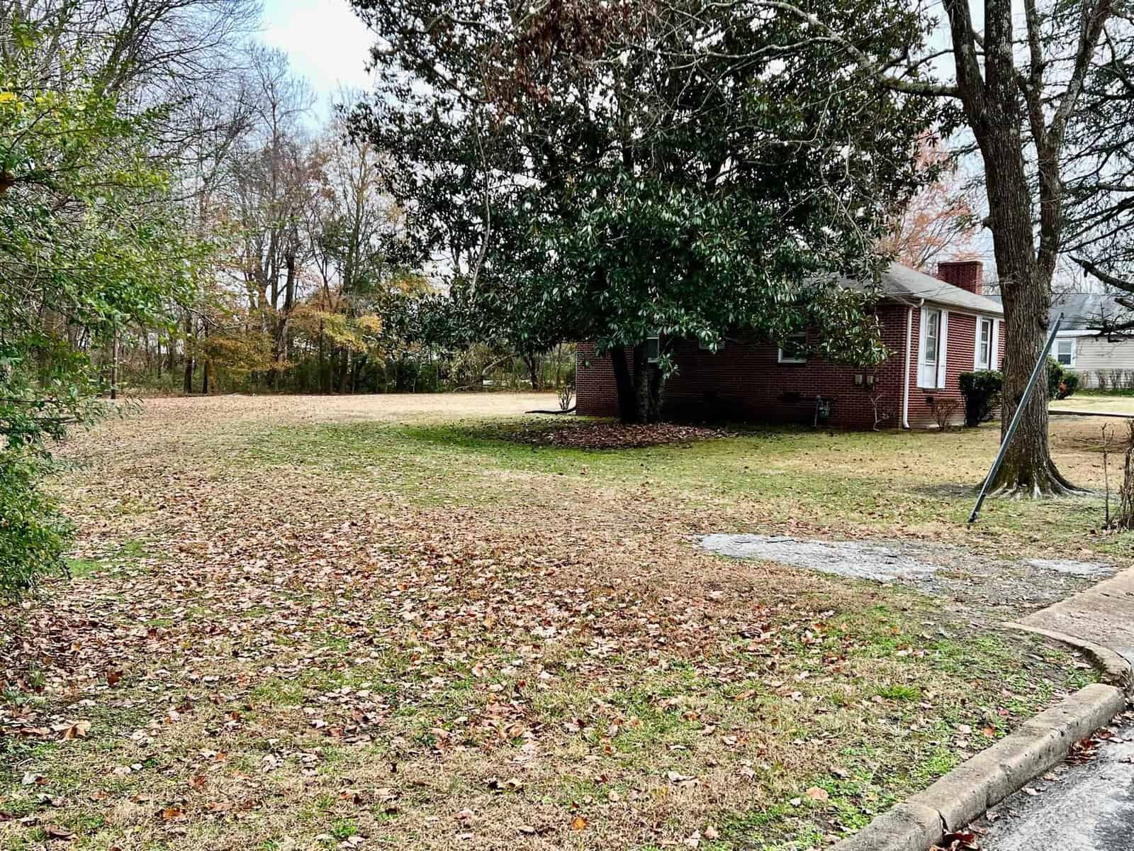 A residential area with a single-story brick house, a front yard covered in fallen leaves, and a tree on the left-hand side, with overcast skies above.