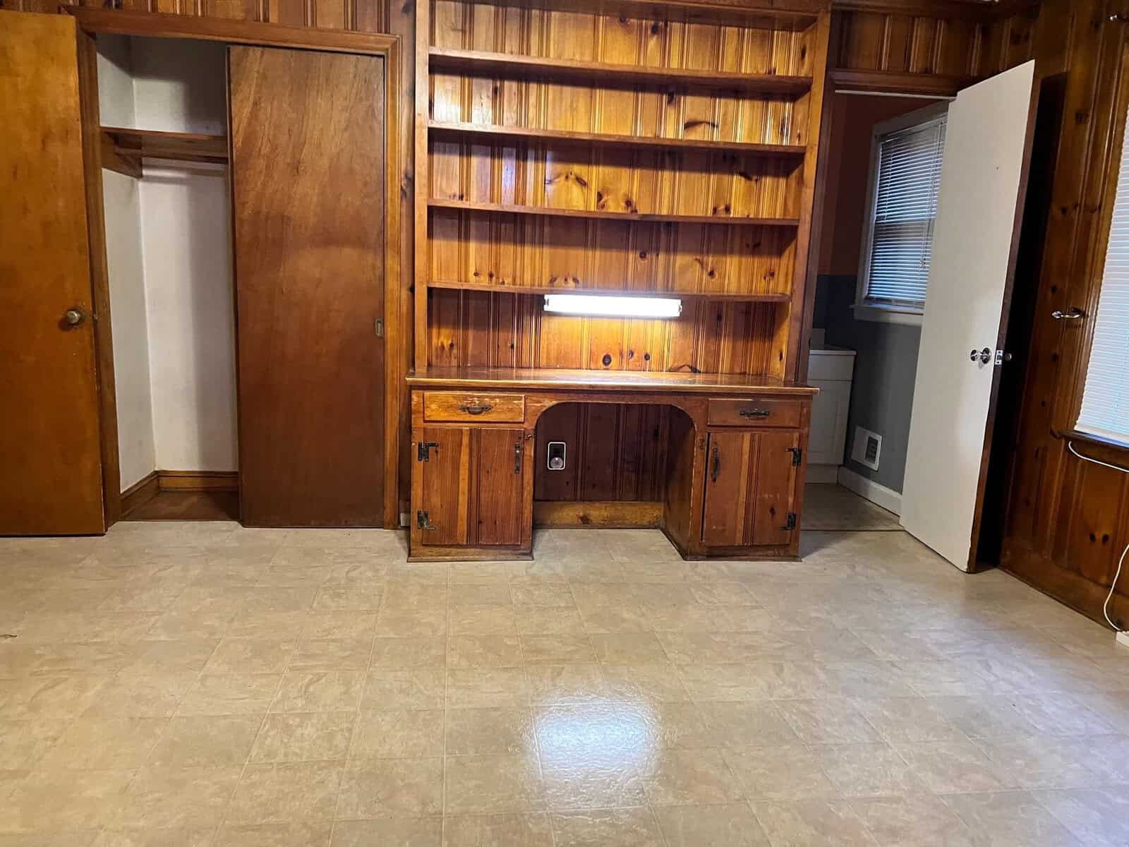 A vacant room with a built-in wooden desk and shelving unit, featuring linoleum flooring and a reflection from the overhead lighting.
