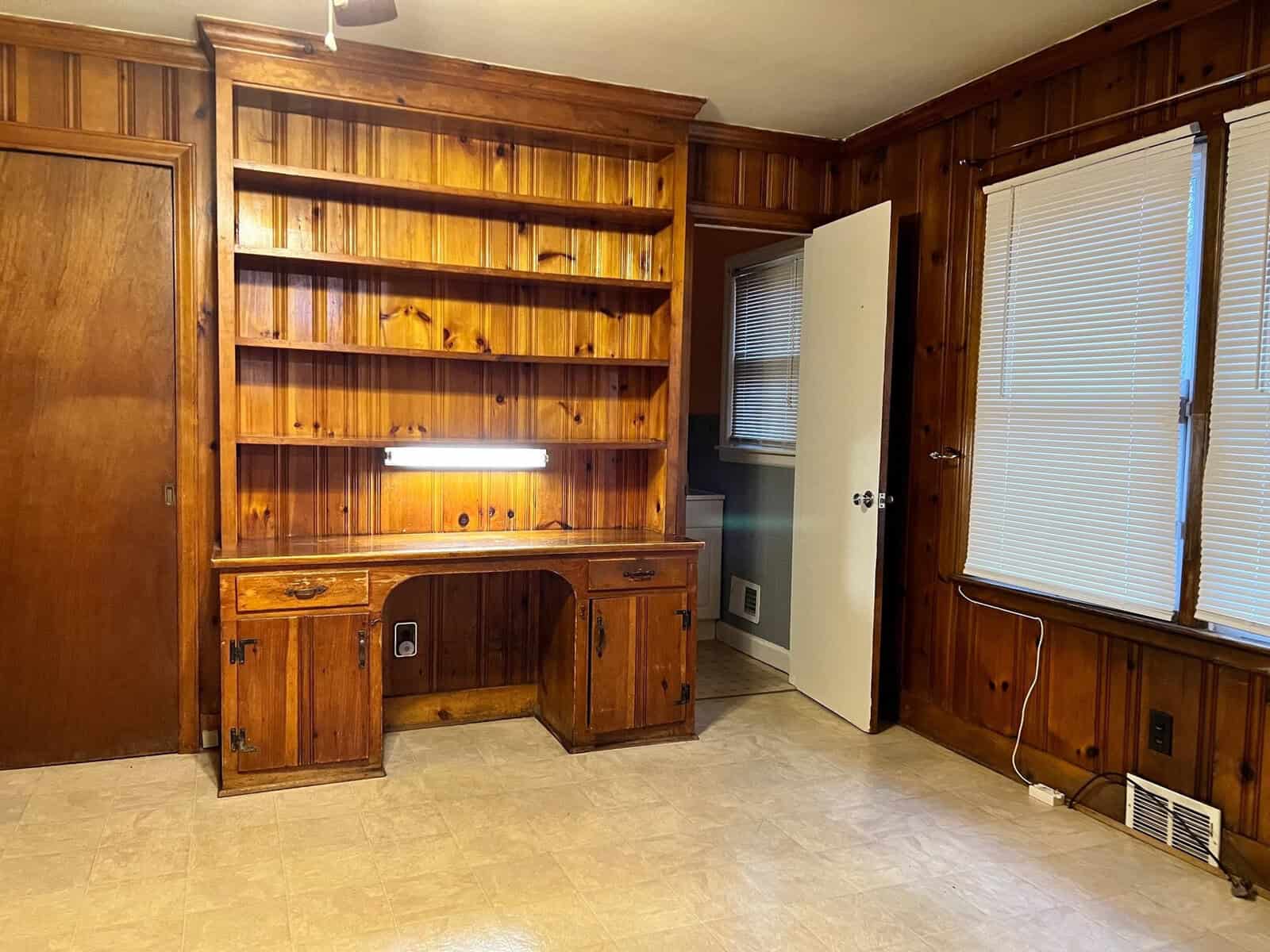 An empty room featuring wood-paneled walls, a built-in wooden desk with shelves, and a linoleum floor.