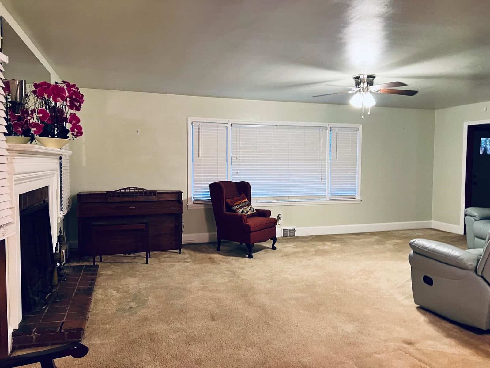 A living room interior featuring a beige carpet, with a piano on the left, a red wingback chair by the window, and a light blue leather sofa on the right. the room has a fireplace on the left and is illuminated by a ceiling fan light fixture.