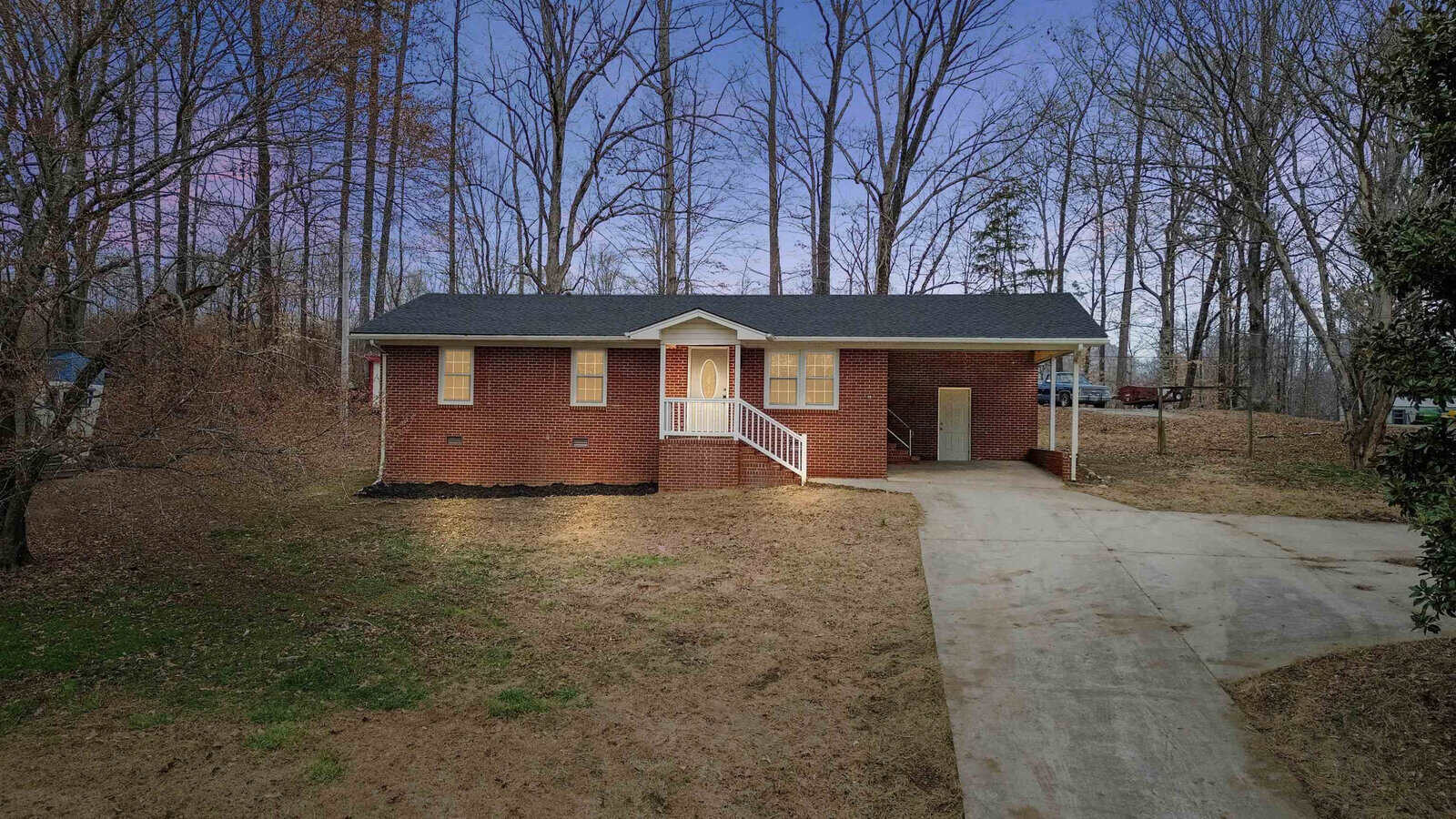 A brick, single-story house with a front porch and white railings is nestled among bare trees on 820 Egypt Mountain Road in Kittrell. There is a driveway leading to a carport on the right side of the house. The large yard has patches of grass among the barren ground, and the sky is clear.