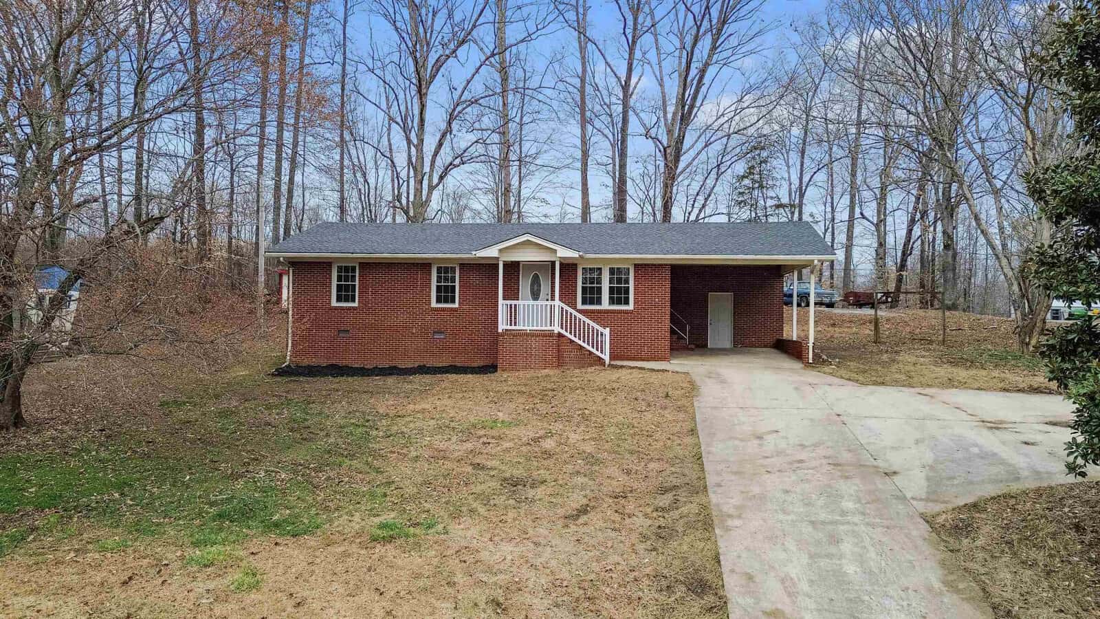 A single-story red-brick house stands at the end of a driveway surrounded by bare trees. The home has a dark shingled roof, a white front door, and a small porch with white railing. A carport is attached to the right side of the house. The yard is mostly bare with patches of grass.